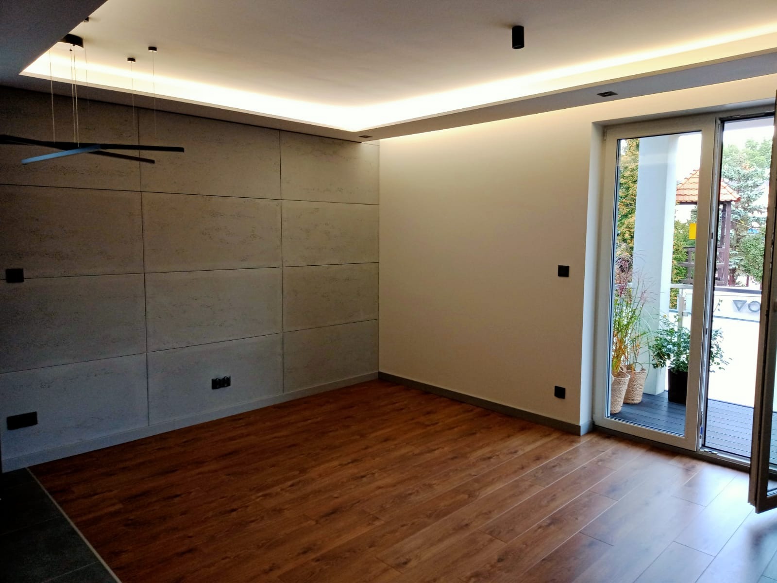 Nowoczesne wnętrze z panelami ściennymi imitującymi beton, drewnianą podłogą i oświetleniem LED w suficie podwieszanym, widok na balkon przez otwarte drzwi balkonowe.