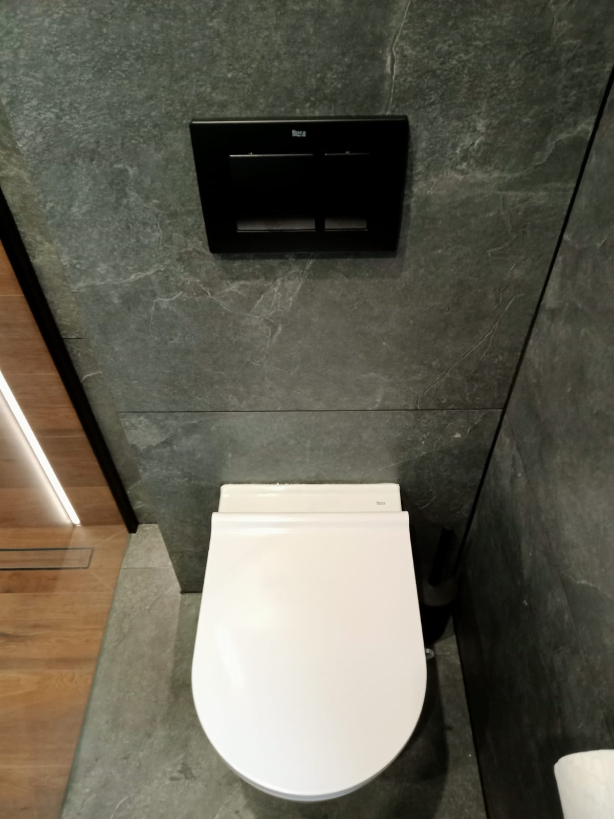 Nowoczesna, podwieszana toaleta w łazience wykończonej szarym kamieniem, z czarnym panelem spłukującym Roca i odpływem liniowym w podłodze.