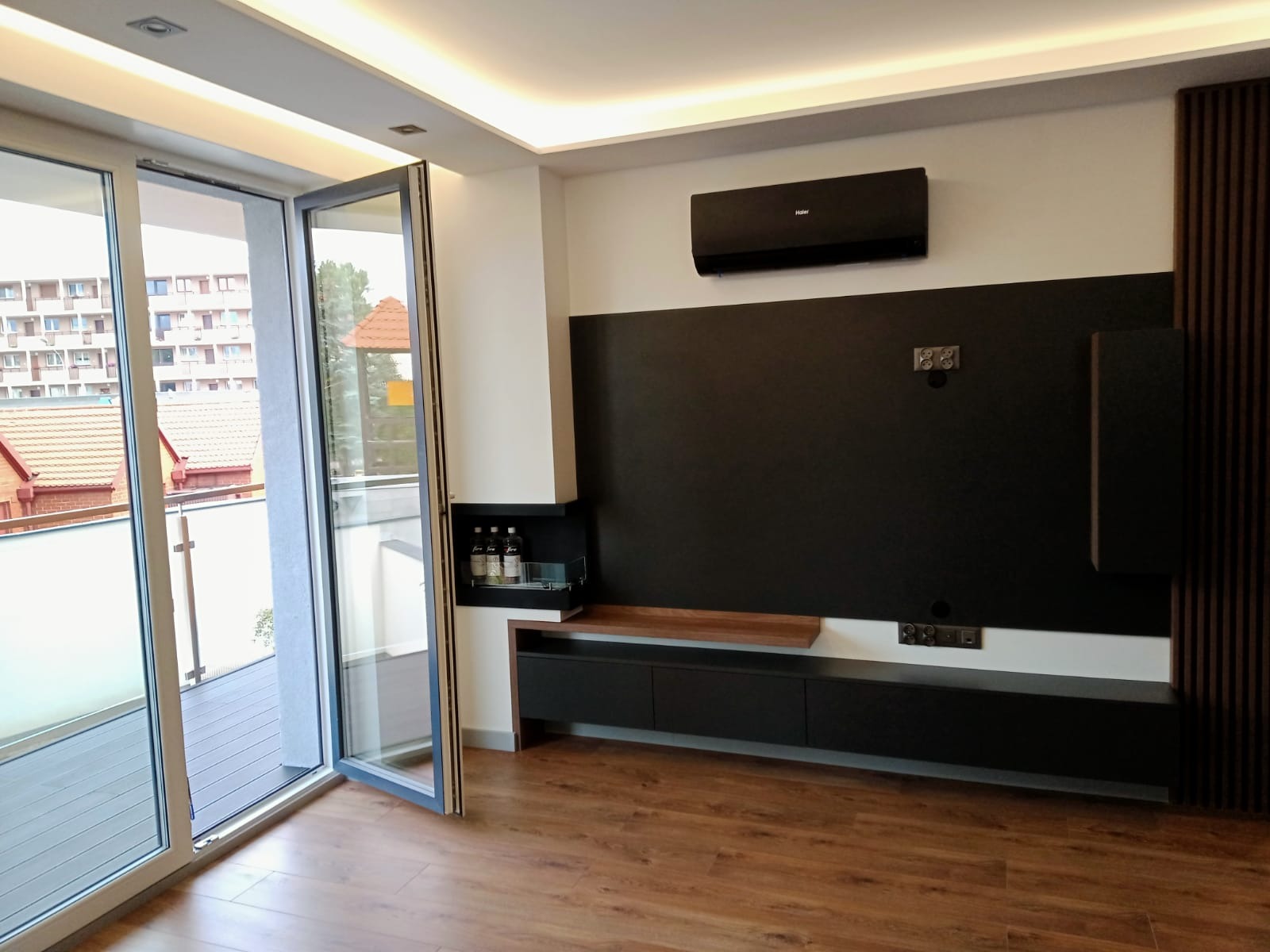 Nowoczesny salon z dużym oknem balkonowym, czarną ścianą TV z szafką RTV, klimatyzatorem Haier i drewnianą podłogą.