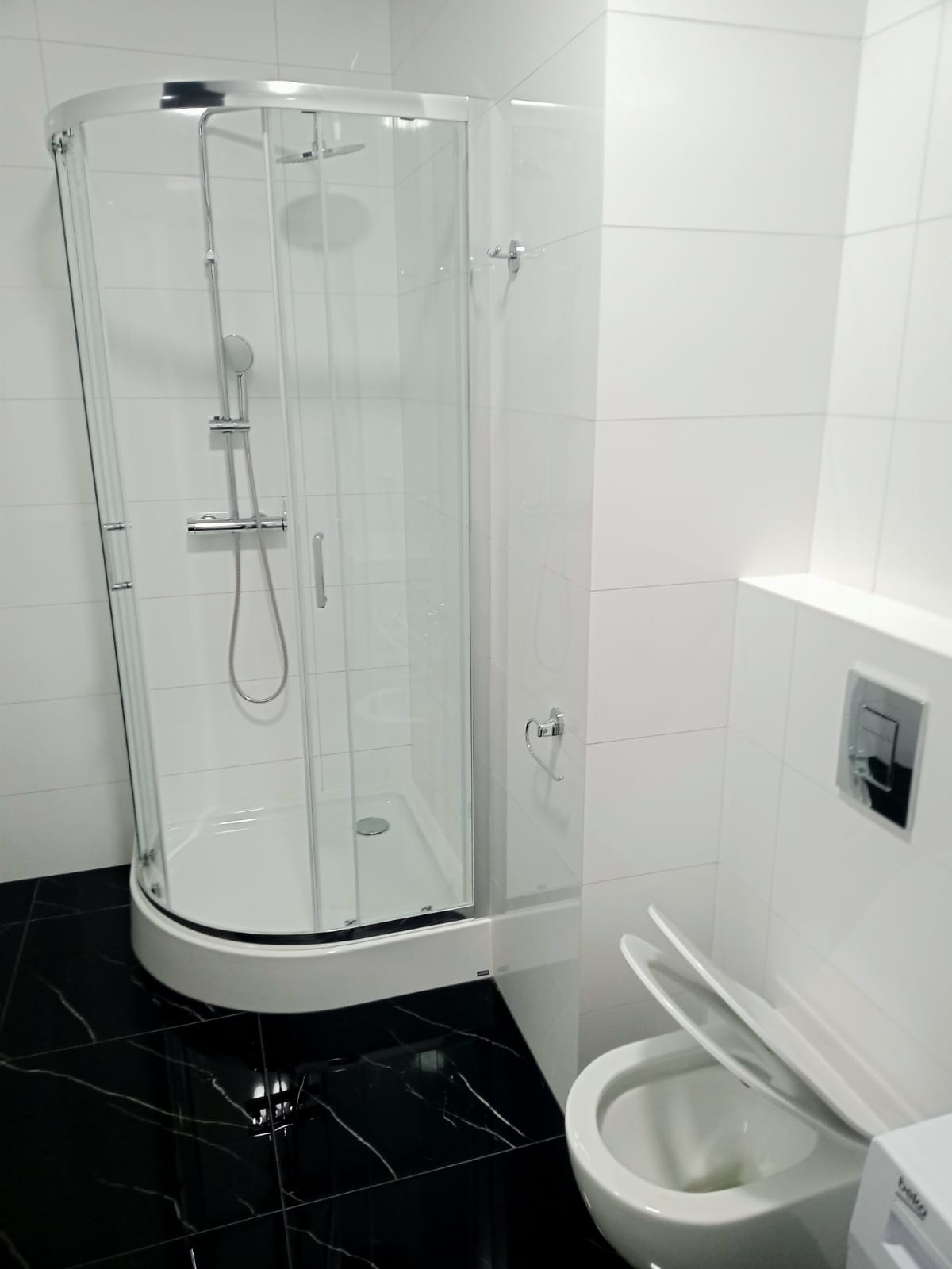 Nowoczesna łazienka z kabiną prysznicową z chromowanymi detalami, białą ceramiką i czarną, błyszczącą podłogą z białymi żyłkami, widok na częściowo otwartą klapę toalety.