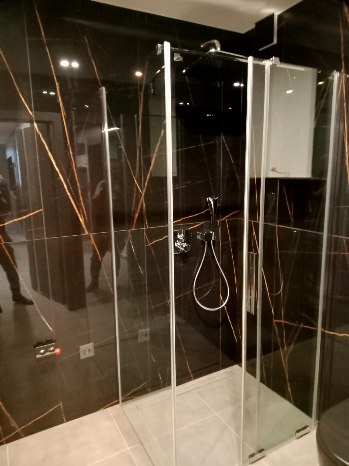 Nowoczesna kabina prysznicowa z czarnymi płytkami imitującymi marmur z złotymi żyłami, szklane drzwi, chromowana armatura, jasna podłoga.