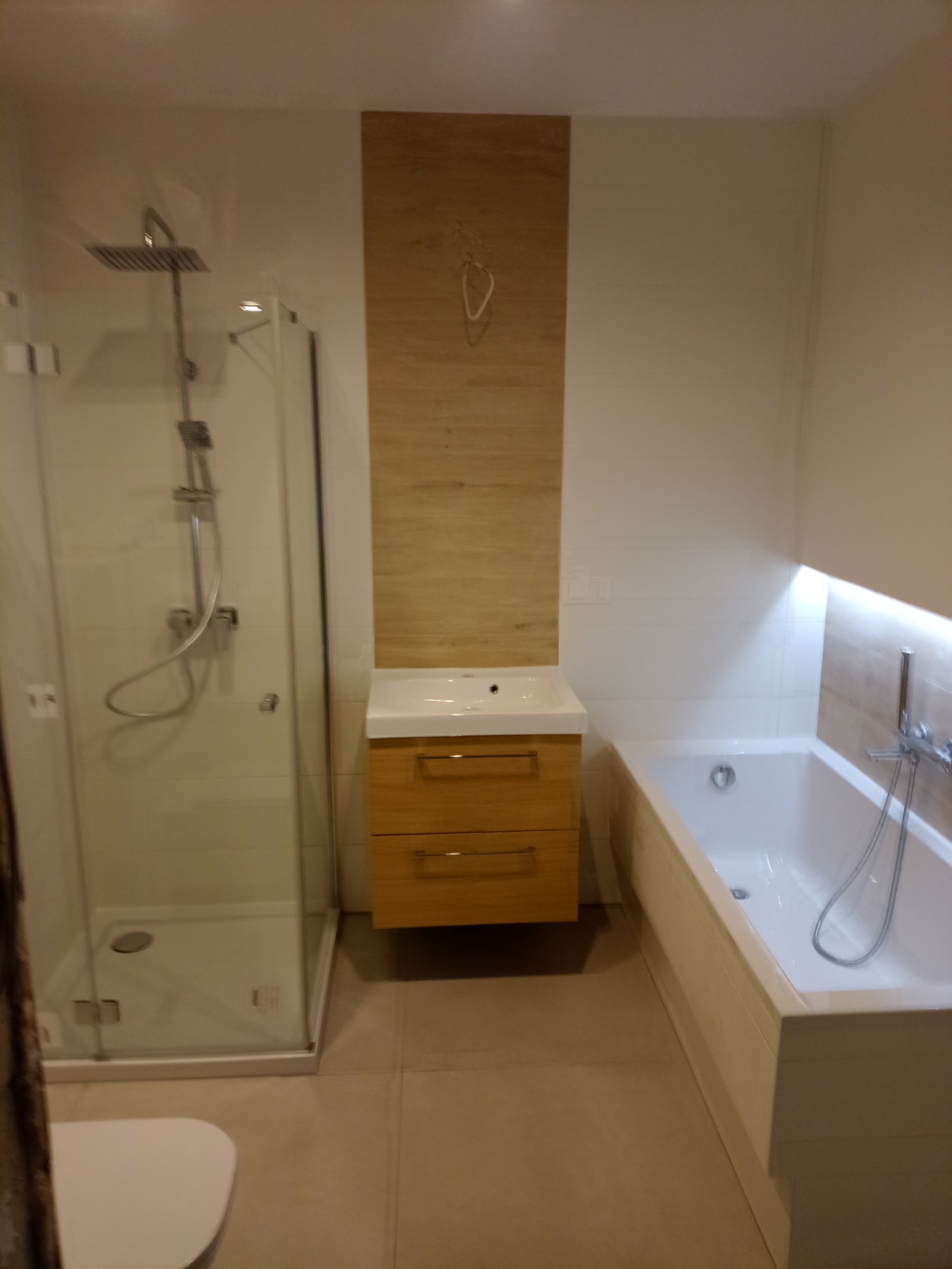 Nowoczesna łazienka z kabiną prysznicową typu walk-in, podświetlaną wanną i szafką z umywalką z drewnianym frontem, wykończona w jasnych, neutralnych kolorach.