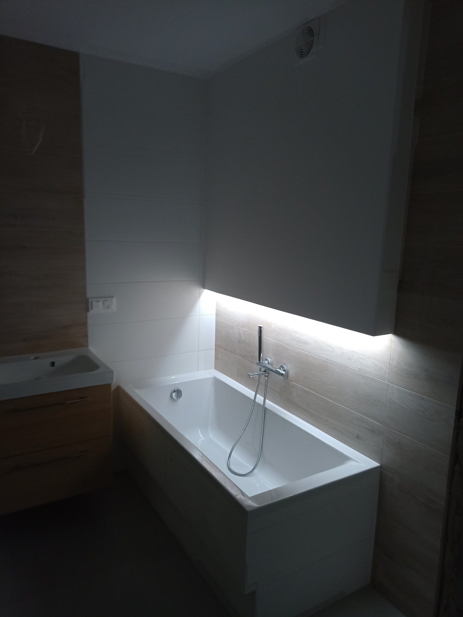 Nowoczesna, biała wanna z chromowaną baterią i wężem prysznicowym, podświetlona od dołu, na tle płytek imitujących drewno, obok umywalka z szafką w podobnym stylu.