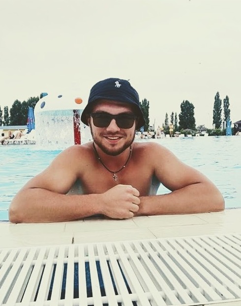 Mężczyzna w okularach przeciwsłonecznych i kapeluszu typu bucket hat opiera się o brzeg basenu w Opolu, w tle widoczny park wodny z fontanną.