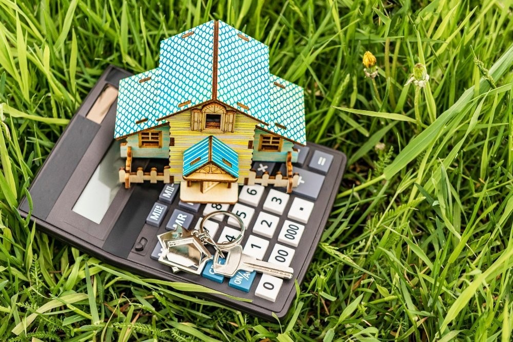 Drewniany model domu z niebieskim dachem i kluczami leżący na kalkulatorze, całość na tle zielonej trawy.