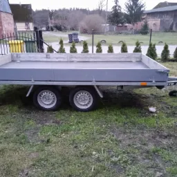 Szara, pusta przyczepa dwuosiowa na trawiastym terenie, z widocznymi budynkami gospodarczymi w tle i rzędem małych drzewek.