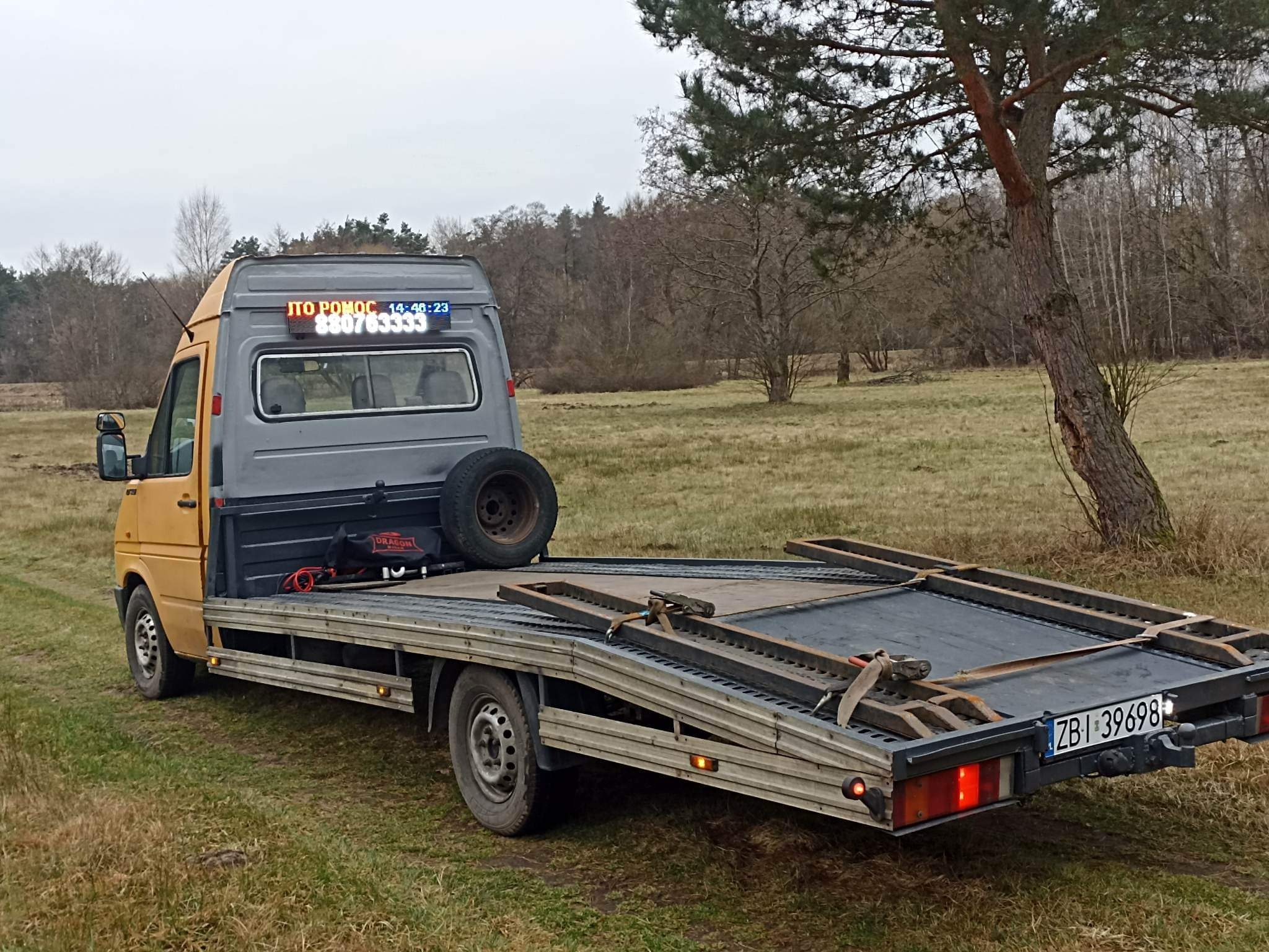 Żółto-szara laweta z wyświetlaczem LED 'ITO POMOC' i numerem telefonu zaparkowana na łące z drzewami w tle.