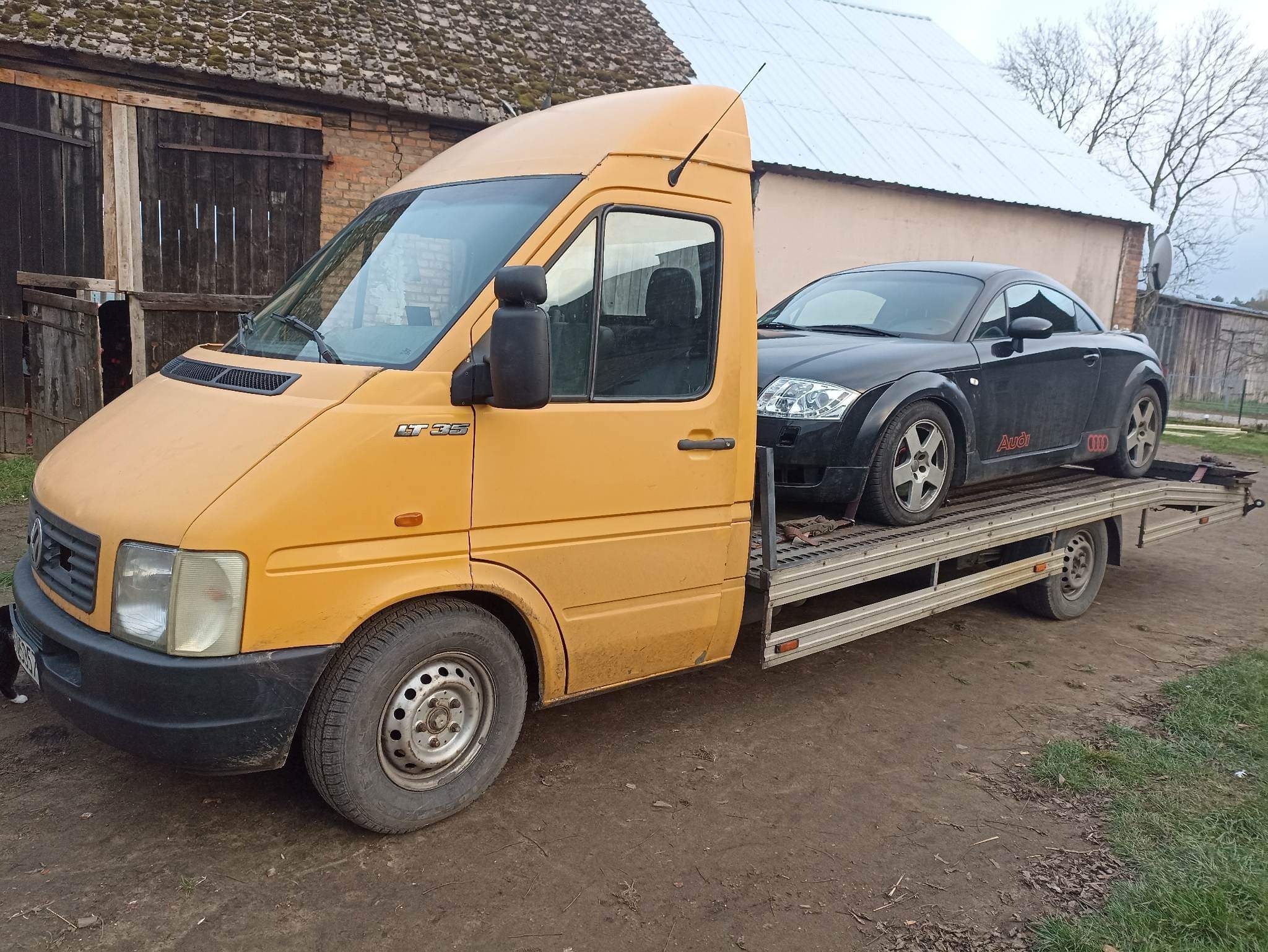 Żółta laweta Volkswagen LT 35 z czarnym Audi TT na platformie, zaparkowana na ziemistej nawierzchni przed budynkami gospodarczymi.