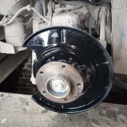 Widok na piastę koła samochodowego z nowo pomalowaną na czarno osłoną tarczy hamulcowej, widoczne elementy zawieszenia i sprężyna amortyzatora, tło stanowi ceglana ściana.