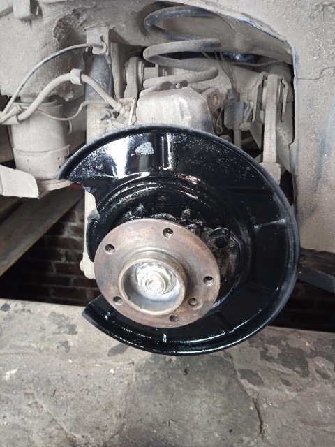 Widok na piastę koła samochodowego z nowo pomalowaną na czarno osłoną tarczy hamulcowej, widoczne elementy zawieszenia i sprężyna amortyzatora, tło stanowi ceglana ściana.