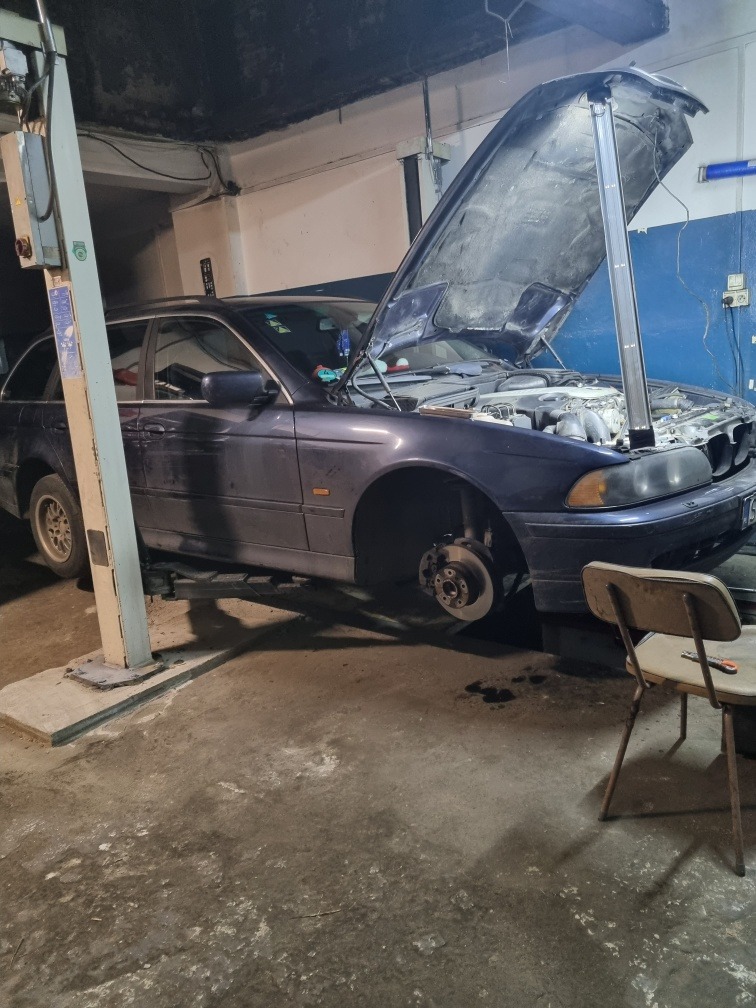 Niebieski samochód kombi na podnośniku w warsztacie z otwartą maską i zdjętym kołem, widoczny silnik, obok krzesło i narzędzie.