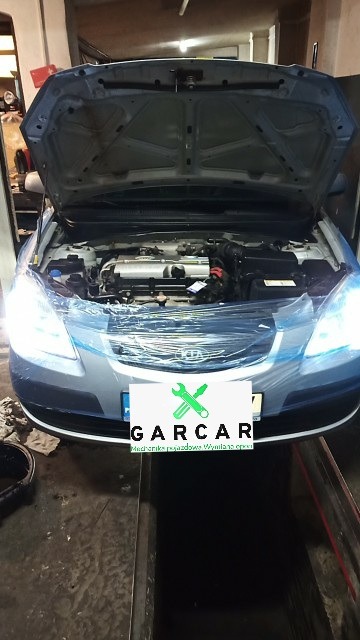 Srebrny samochód z otwartą maską w warsztacie, silnik widoczny, przód auta zabezpieczony folią ochronną, w tle widoczna kanalizacja warsztatowa.