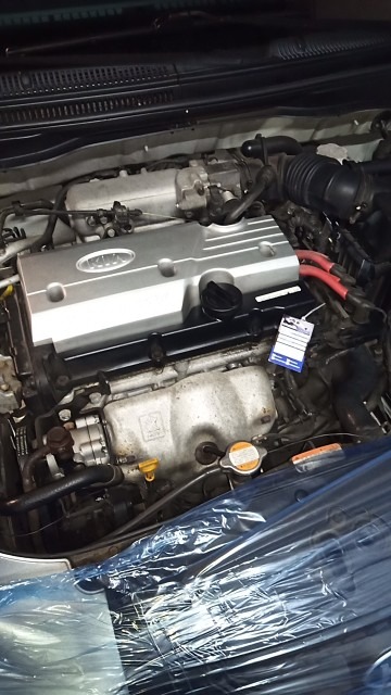 Silnik samochodu marki KIA z widoczną pokrywą z logo, częściowo osłonięty folią ochronną, z zawieszoną kartką informacyjną.