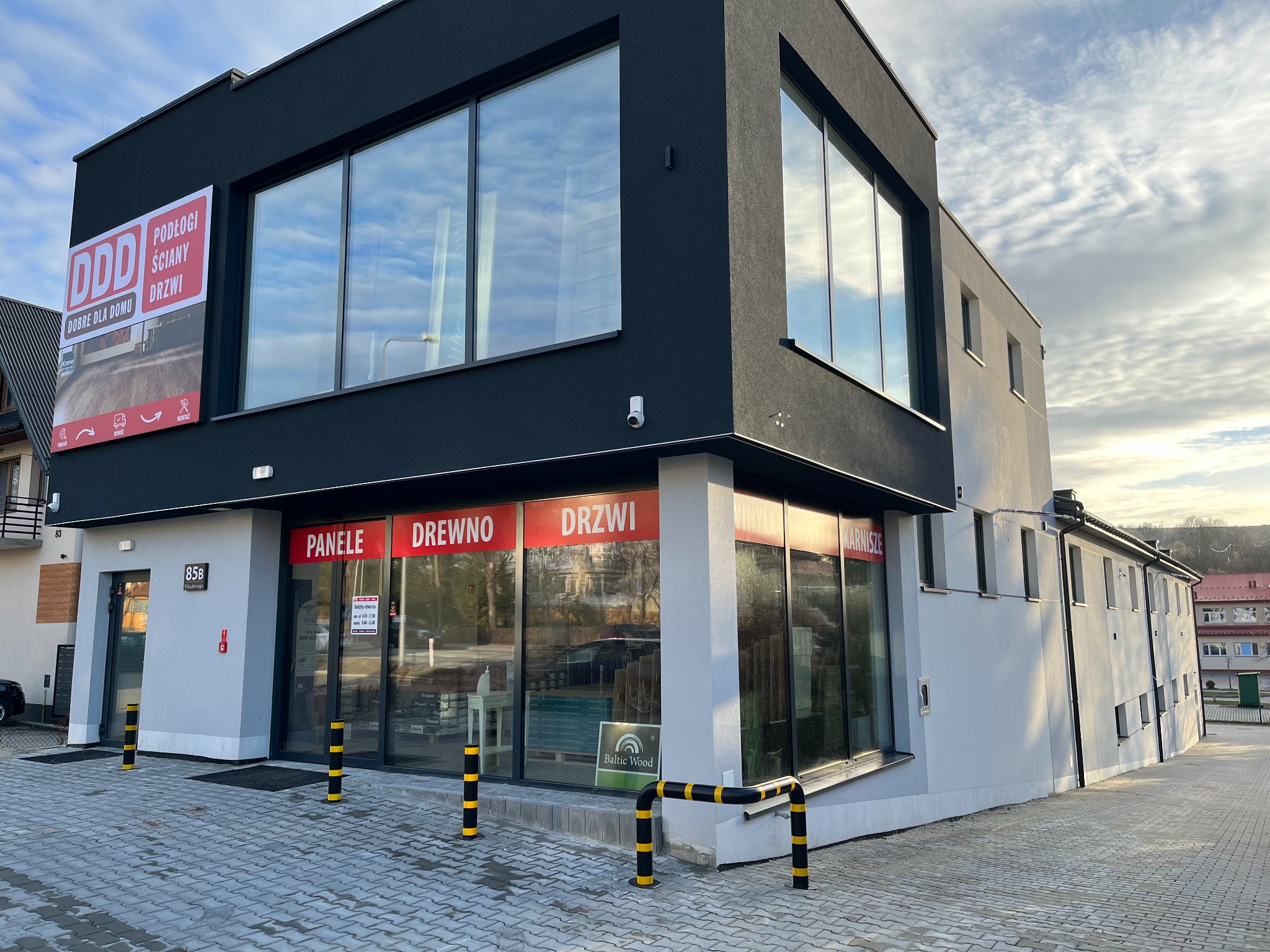 Fasada budynku salonu sprzedaży podłóg, ścian i drzwi z dużymi witrynami, na których widnieją napisy 'Panele', 'Drewno', 'Drzwi', 'Rolety', 'Karnisze', przed wejściem barierki ochronne w kolorze...
