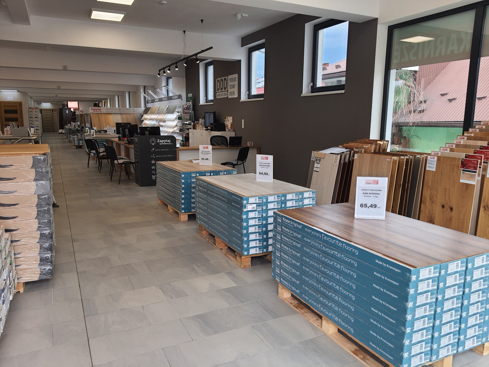 Wnętrze salonu z ekspozycją paneli podłogowych Krono Original na paletach, widoczne ceny i logo producenta, w tle biurka obsługi klienta i próbki podłóg.