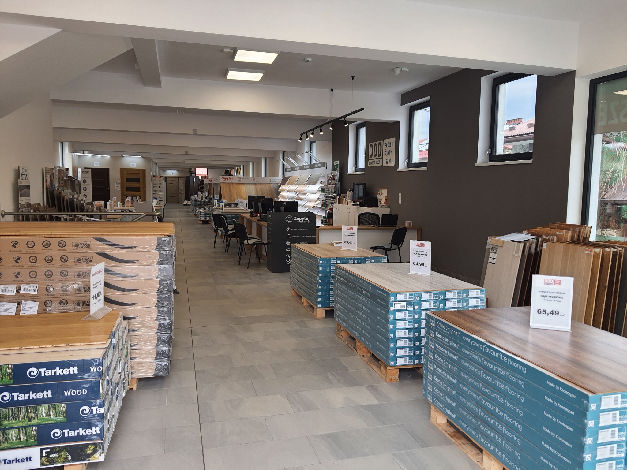 Wnętrze salonu z ekspozycją paneli podłogowych marek Tarkett i Krono Original ułożonych na paletach, widoczne również ekspozytory z próbkami podłóg i drzwi w głębi sklepu.