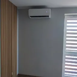 Biały klimatyzator ścienny zamontowany w nowoczesnym salonie z drewnianą ścianą dekoracyjną i żaluzjami okiennymi.