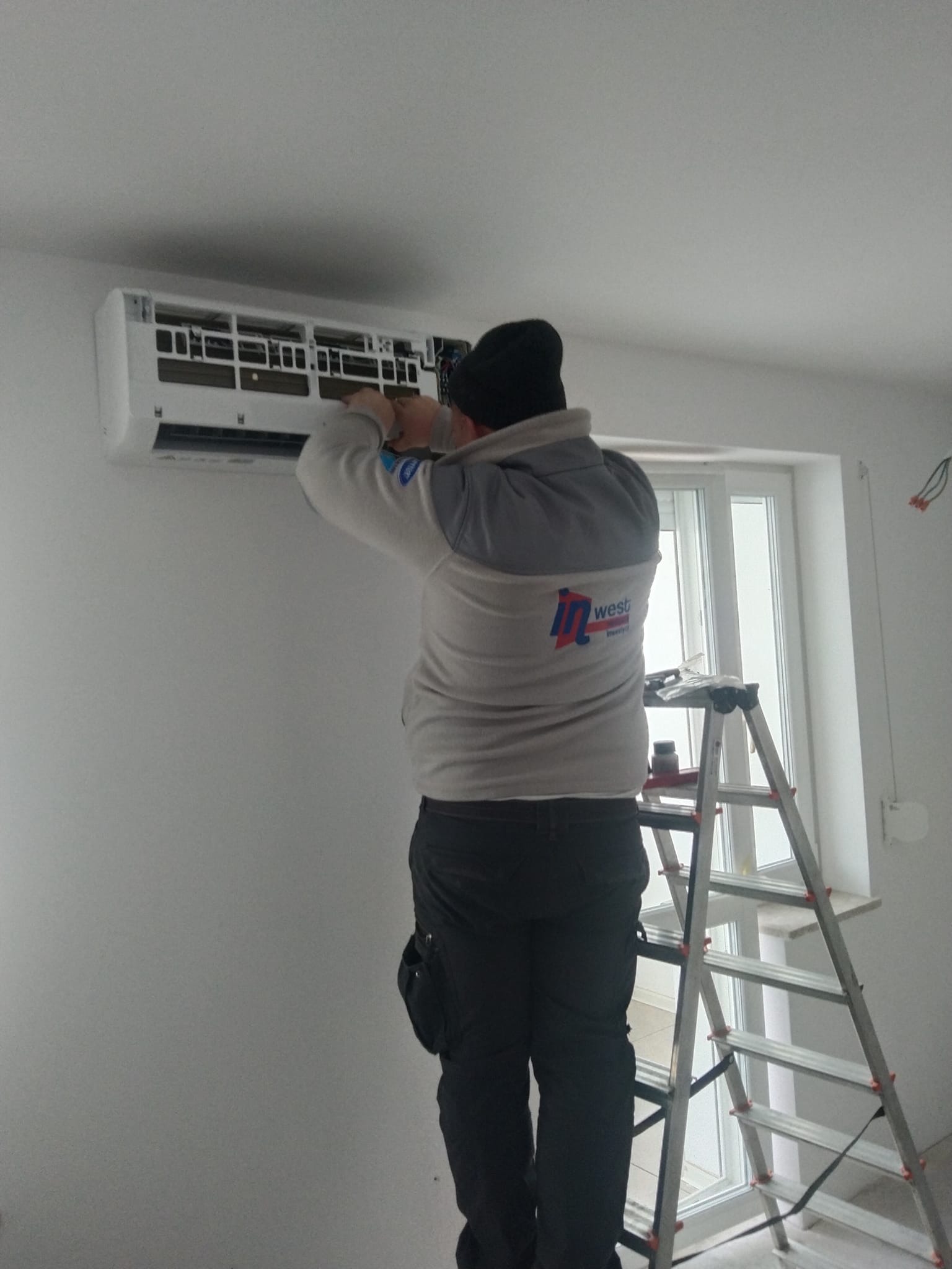 Instalator na drabinie montuje klimatyzator ścienny w jasnym pomieszczeniu, widoczne narzędzia na drabinie, instalacja w trakcie.