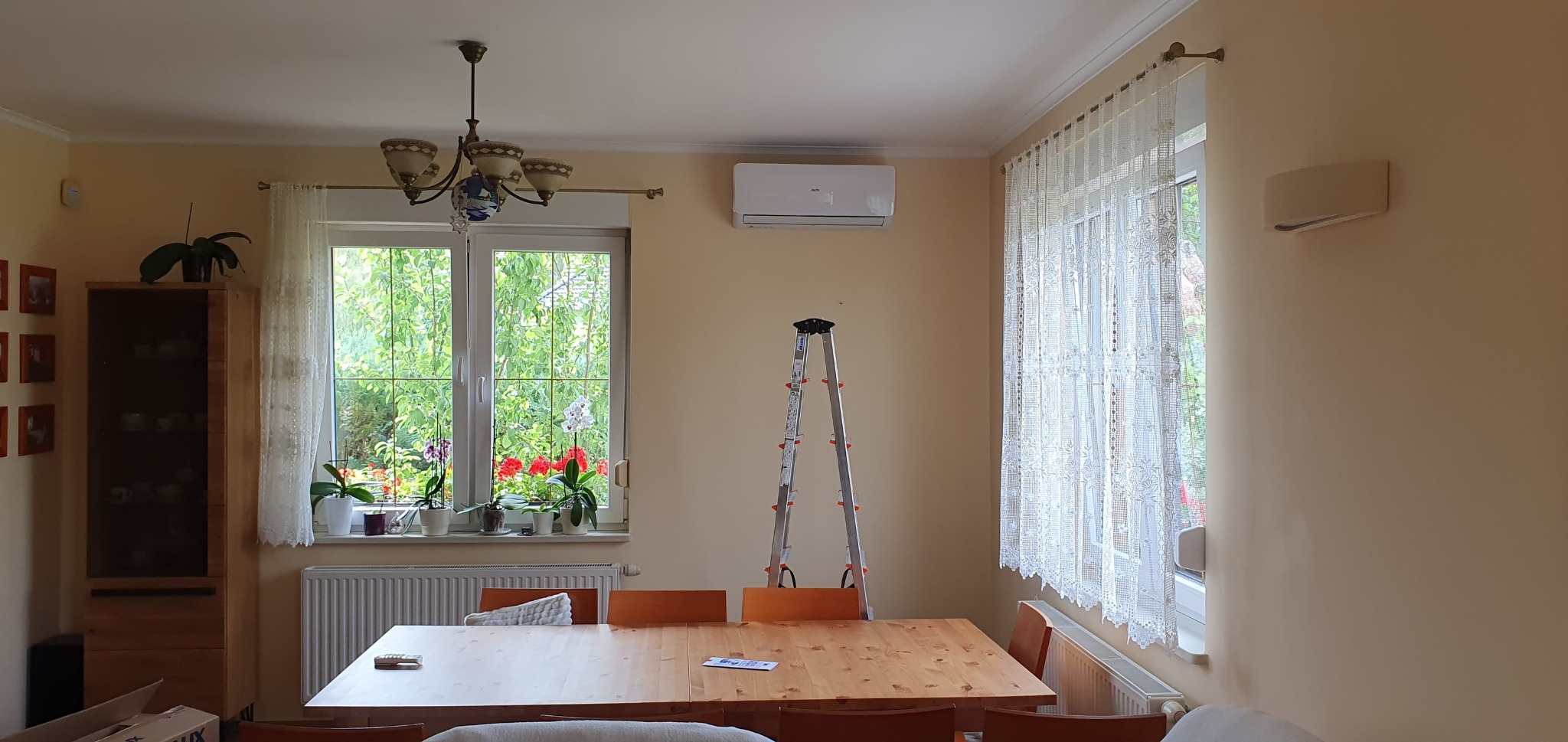 Wnętrze pokoju z zamontowaną klimatyzacją na ścianie, drabina rozstawiona pod klimatyzatorem, okno z kwiatami i firanką w tle.