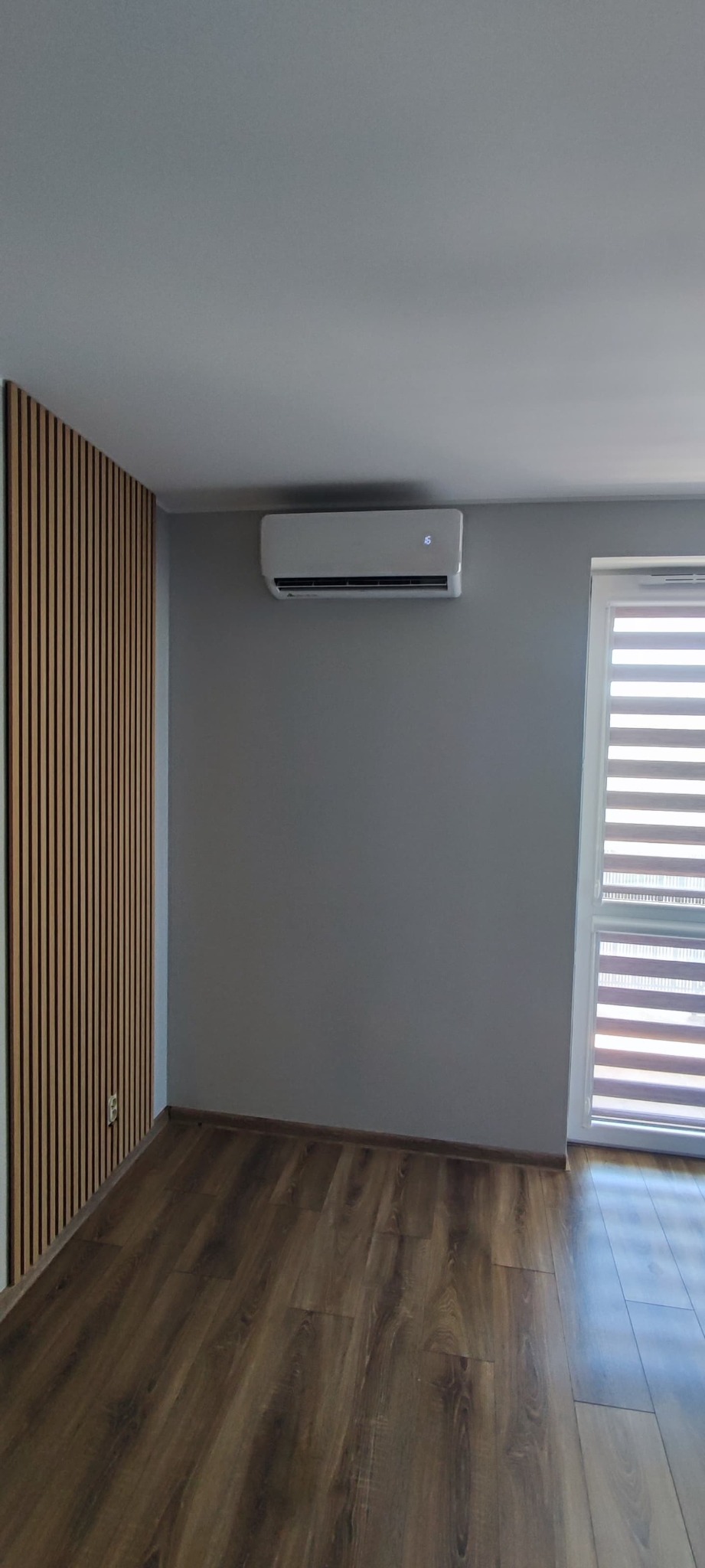 Biały klimatyzator ścienny zamontowany w nowoczesnym salonie z drewnianą ścianą dekoracyjną i żaluzjami okiennymi.