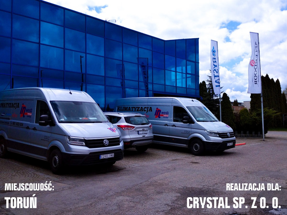 Dwa srebrne busy firmy klimatyzacyjnej zaparkowane przed nowoczesnym biurowcem z niebieską szklaną fasadą, widoczne flagi reklamowe i zaparkowany samochód osobowy pomiędzy nimi.