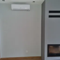 Biała jednostka klimatyzacyjna Hisense zamontowana na szarej ścianie w jasnym pomieszczeniu, widoczny fragment drewnianych drzwi i kominek z półkami.