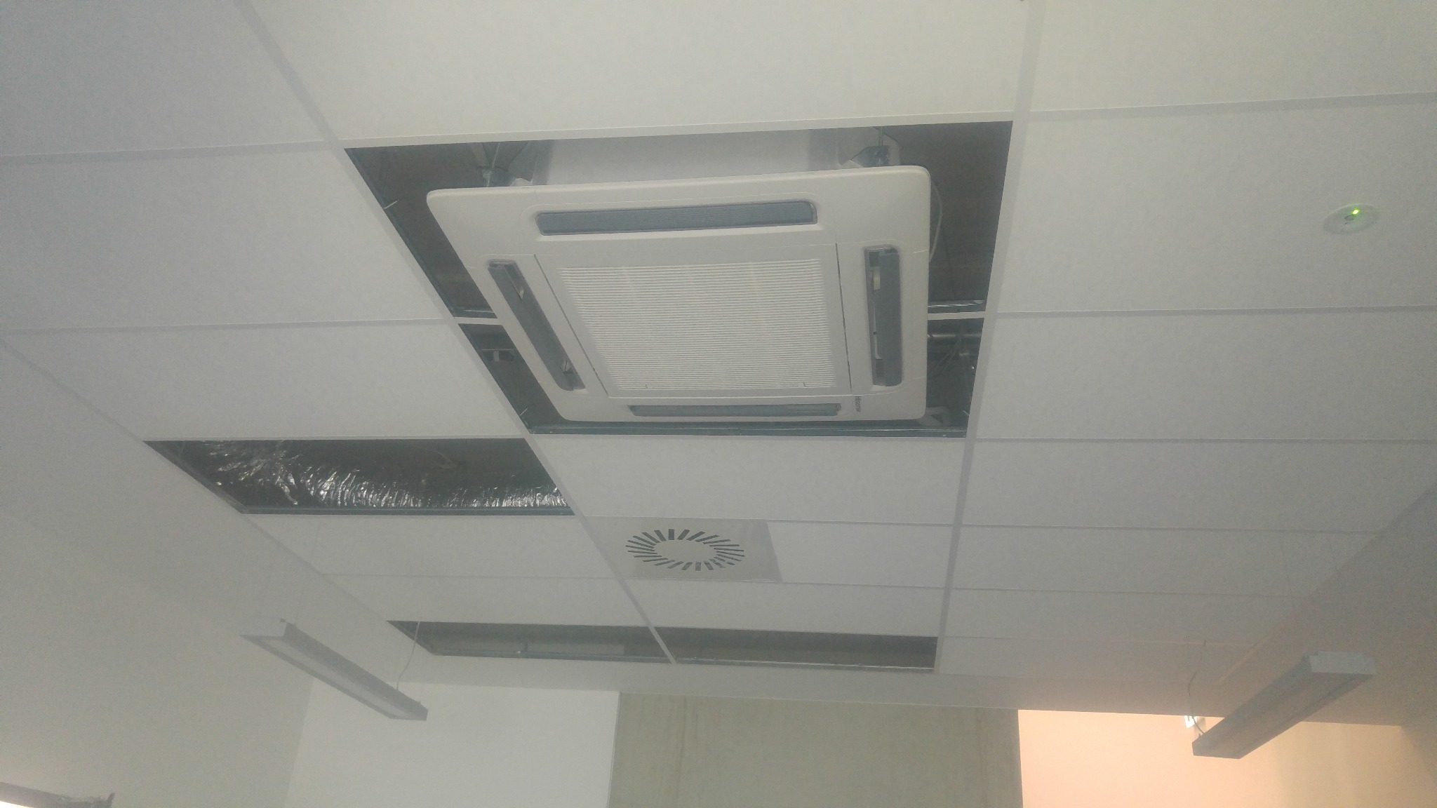 Kasetonowa jednostka klimatyzacji sufitowej, zamontowana w biurze, widoczna w otworze sufitu podwieszanego. Obok widoczny fragment instalacji wentylacyjnej.