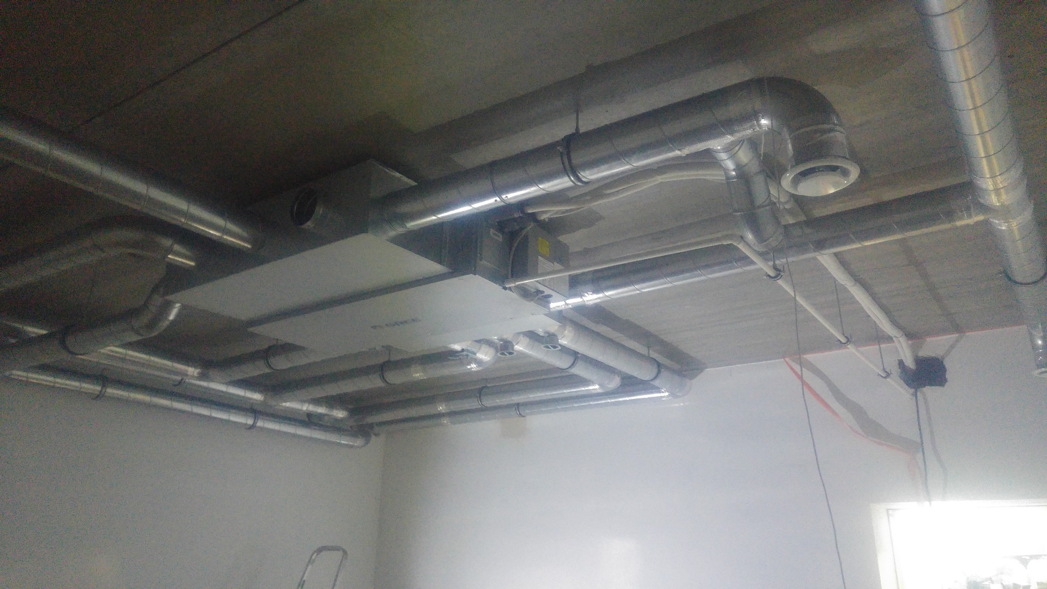 Widok instalacji wentylacyjnej z rurami aluminiowymi pod betonowym sufitem, z widocznymi elementami mocującymi i kratkami wentylacyjnymi, częściowo zakrytej płaskimi panelami.