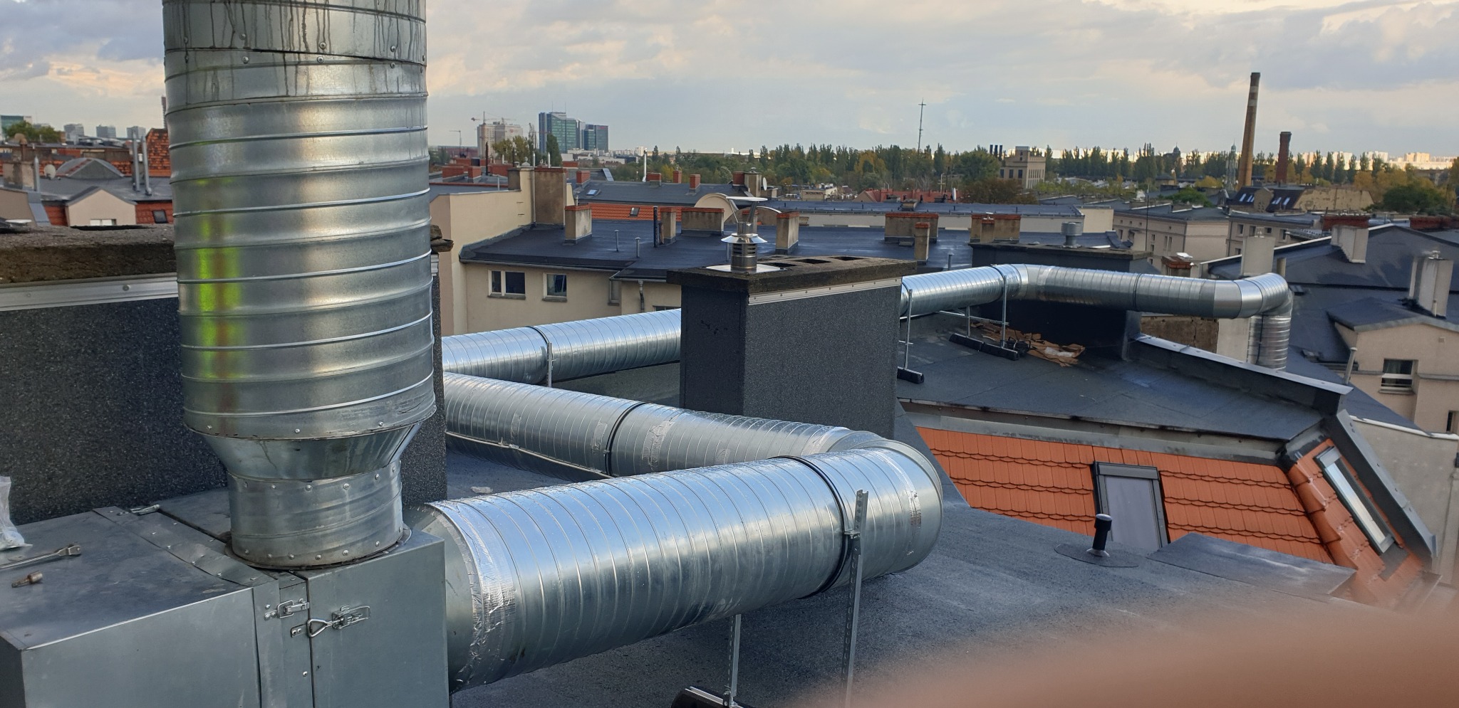 Instalacja wentylacyjna z metalowymi rurami na dachu budynku, widok na panoramę miasta w tle.