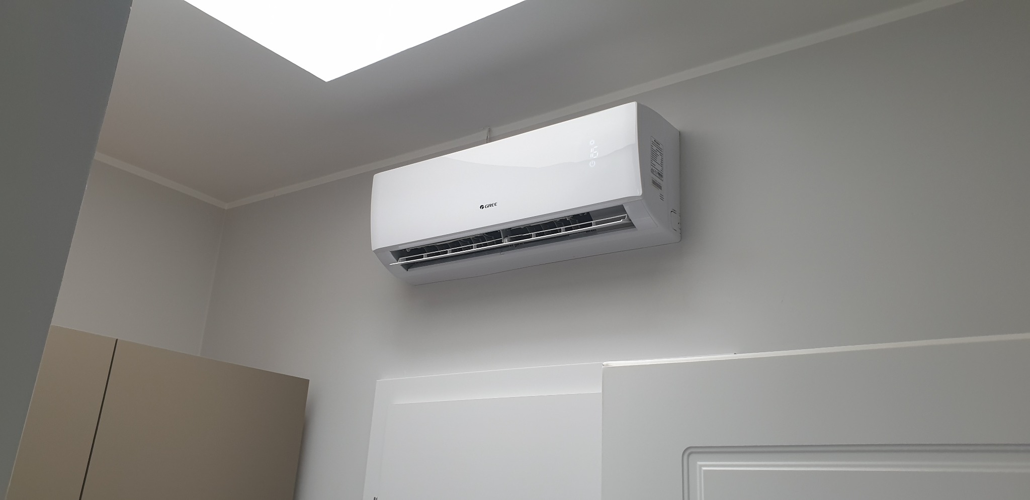 Biały klimatyzator marki Gree zamontowany na jasnoszarej ścianie w pomieszczeniu z widocznym fragmentem szafki i drzwi, oświetlony światłem z góry.