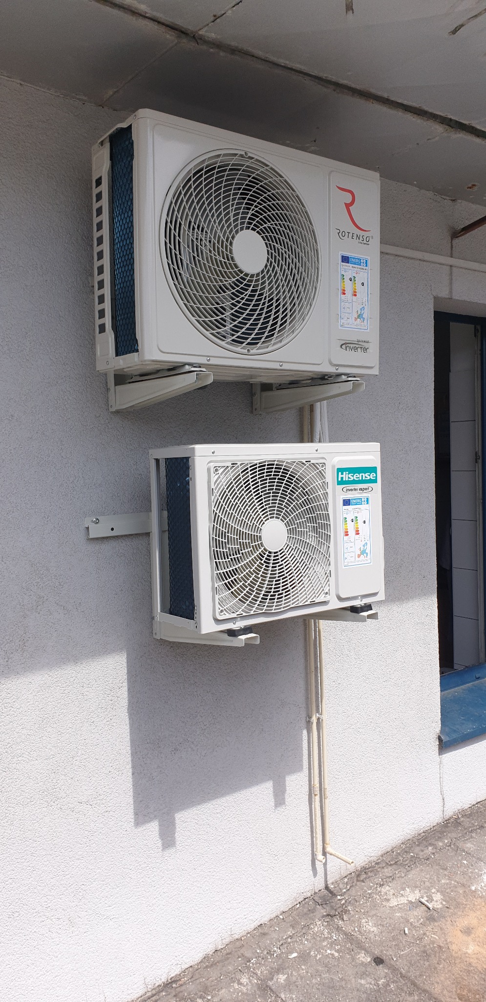 Dwa klimatyzatory różnych marek (Rotenso i Hisense) zamontowane jeden nad drugim na zewnątrz budynku z oknem w tle.