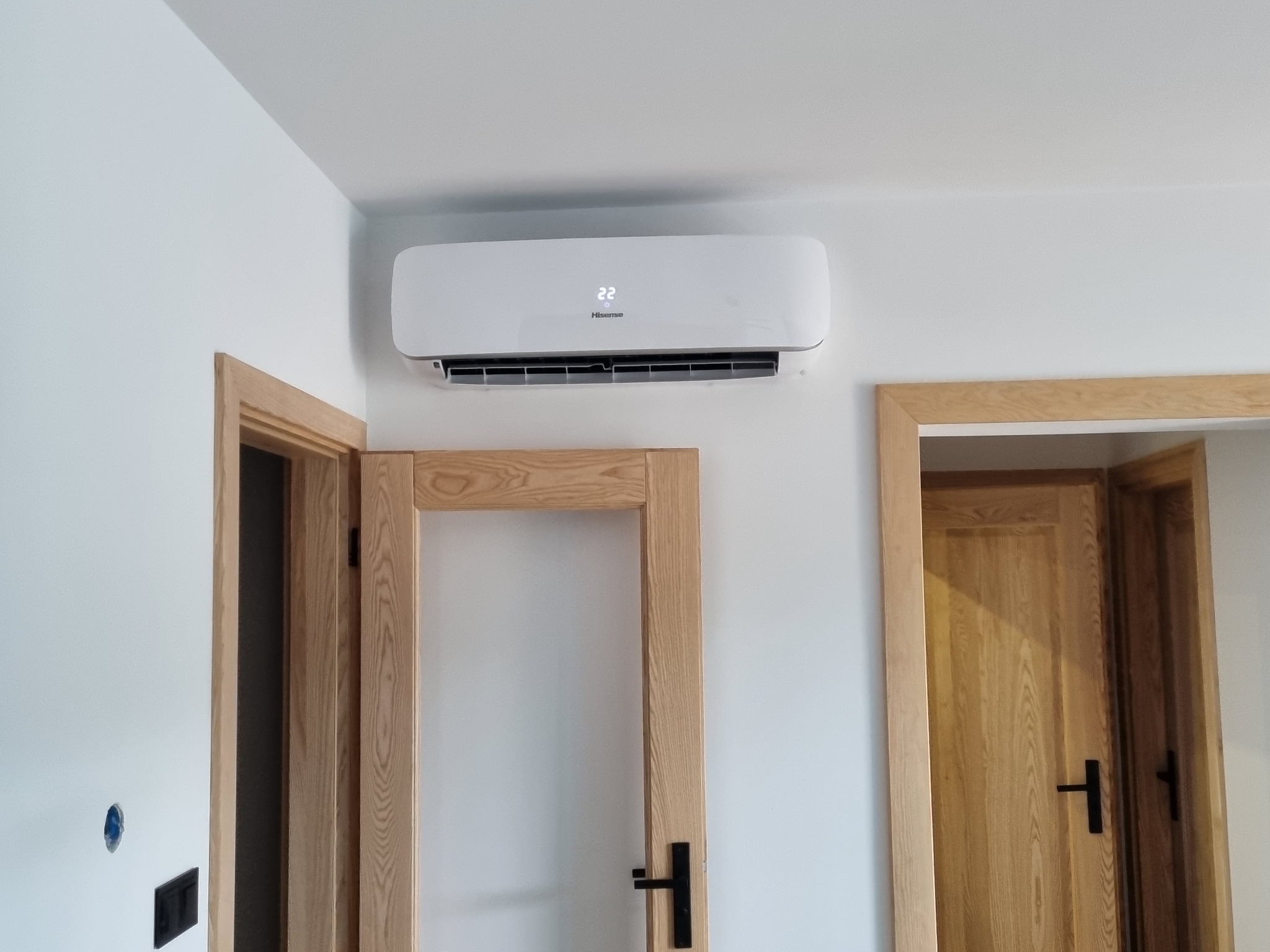 Biały klimatyzator Hisense z wyświetlaną temperaturą 22 stopnie, zamontowany na ścianie w pomieszczeniu z dwiema drewnianymi ościeżnicami drzwiowymi.