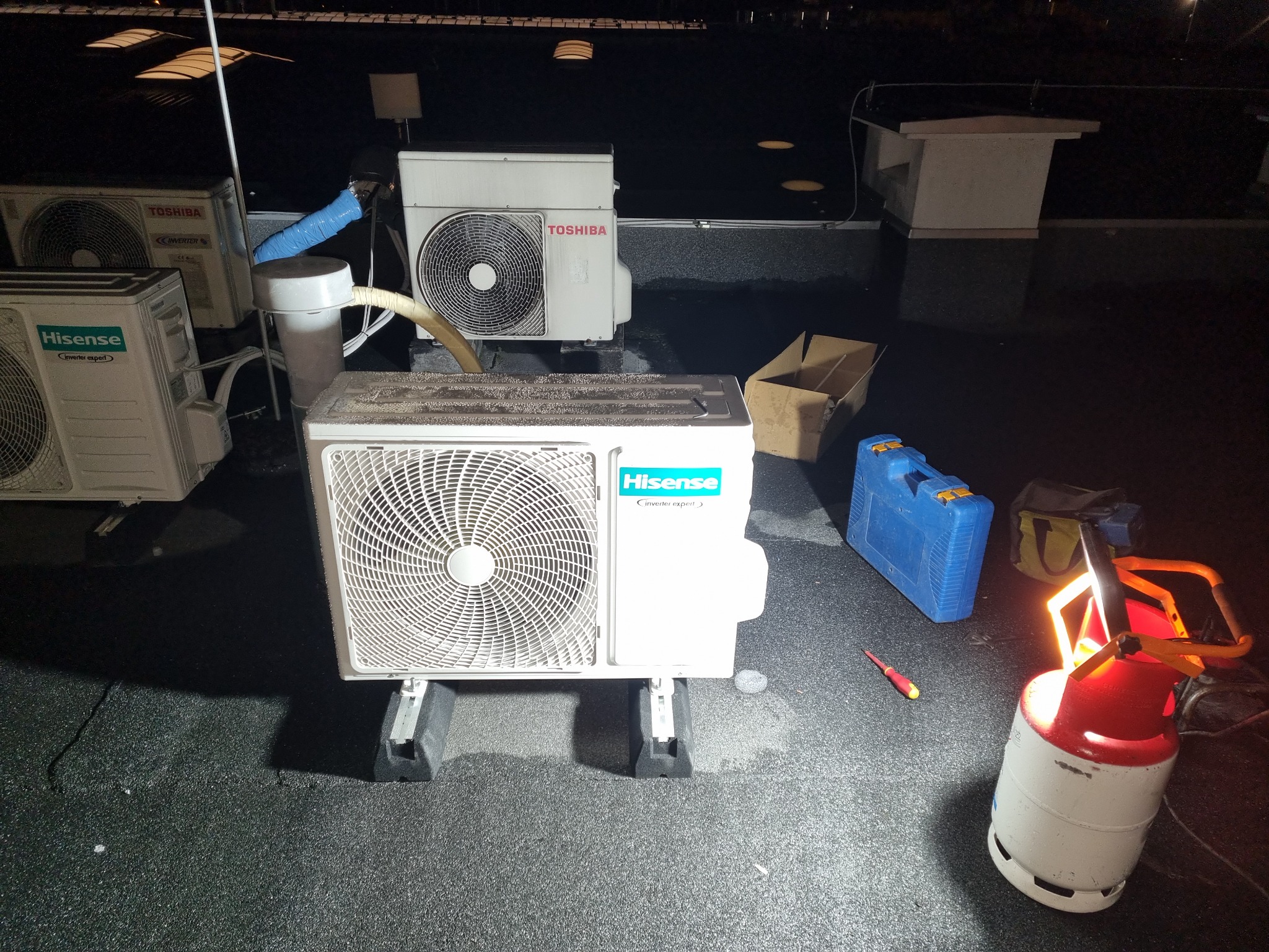 Kilka zewnętrznych jednostek klimatyzacji różnych marek (Hisense, Toshiba) na dachu budynku, obok narzędzia serwisowe, butla z gazem i skrzynka narzędziowa.