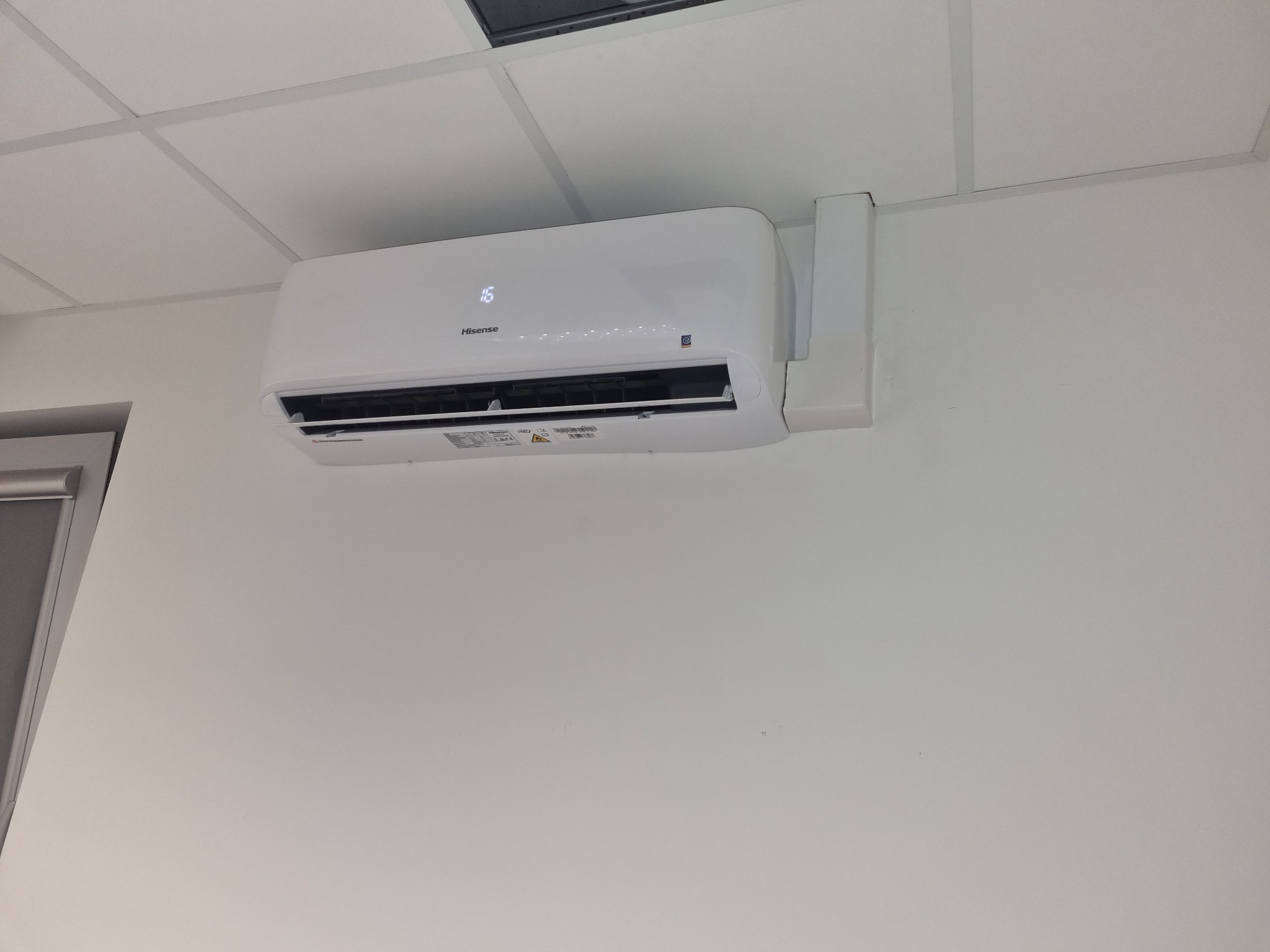 Biała jednostka wewnętrzna klimatyzacji Hisense zamontowana na białej ścianie pod sufitem kasetonowym.