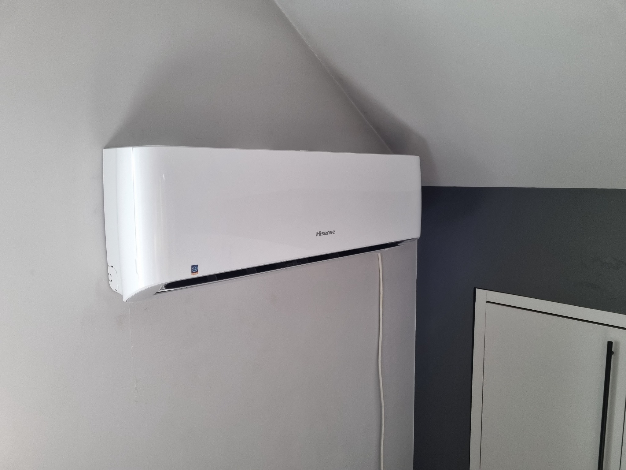 Biała klimatyzacja Hisense zamontowana na szarej ścianie w pokoju z widocznym fragmentem drzwi.