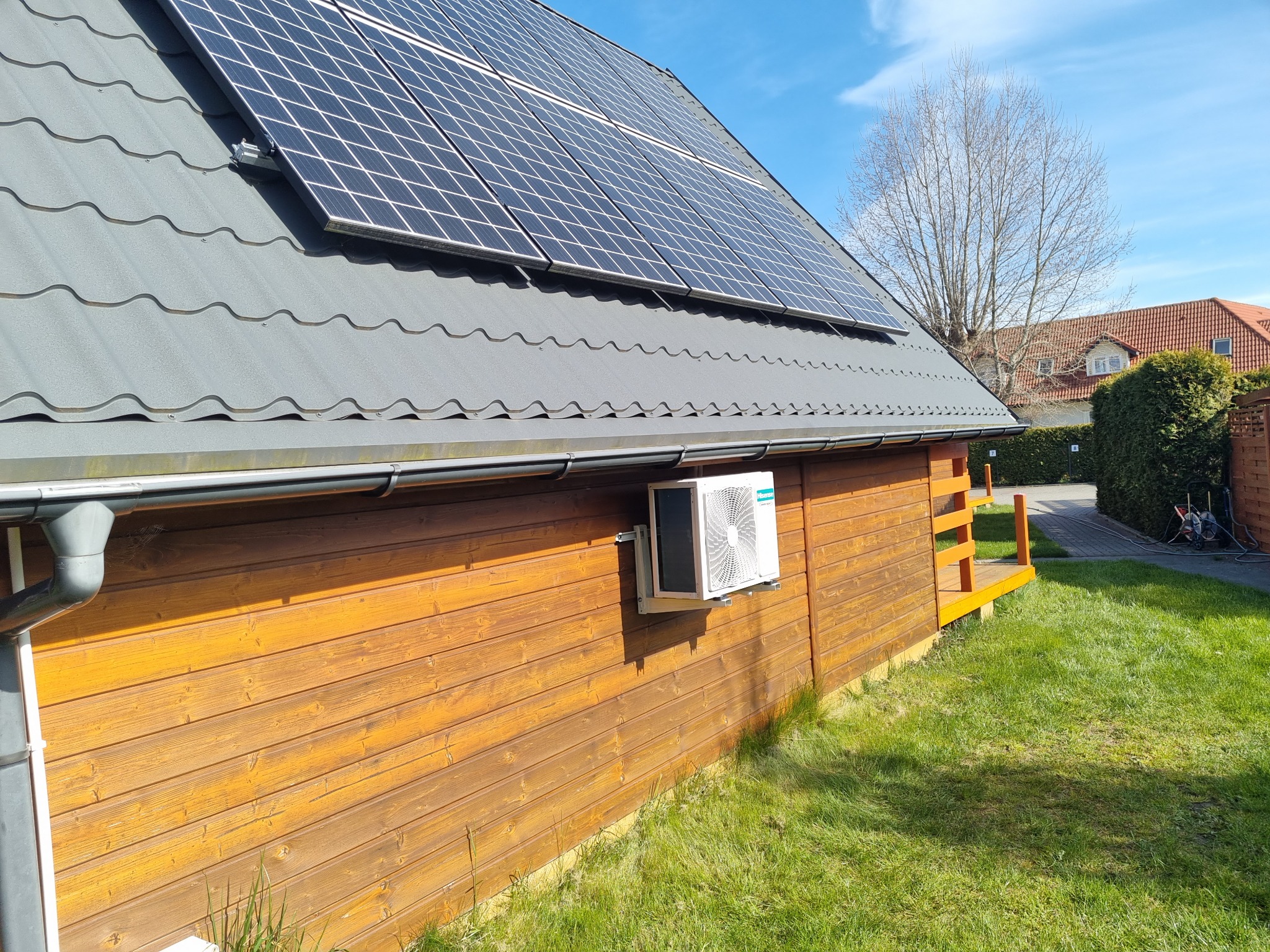 Zewnętrzna jednostka klimatyzacji zamontowana na drewnianej ścianie budynku z panelami słonecznymi na dachu, widok z poziomu trawnika.