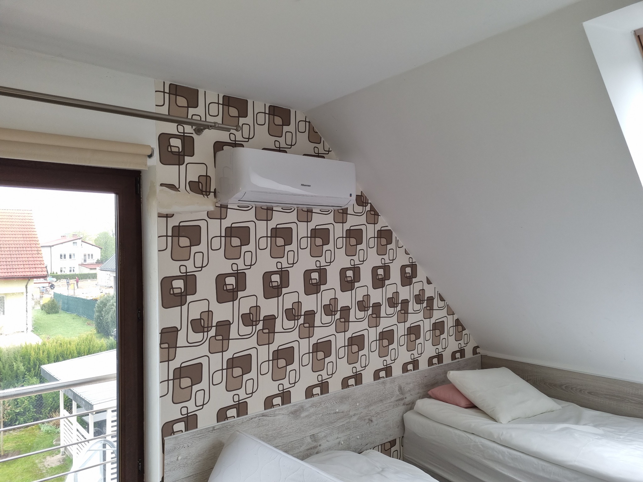 Biała jednostka klimatyzacyjna Hisense zamontowana na ścianie z wzorzystą tapetą w pokoju na poddaszu z widokiem na zewnątrz przez okno balkonowe.