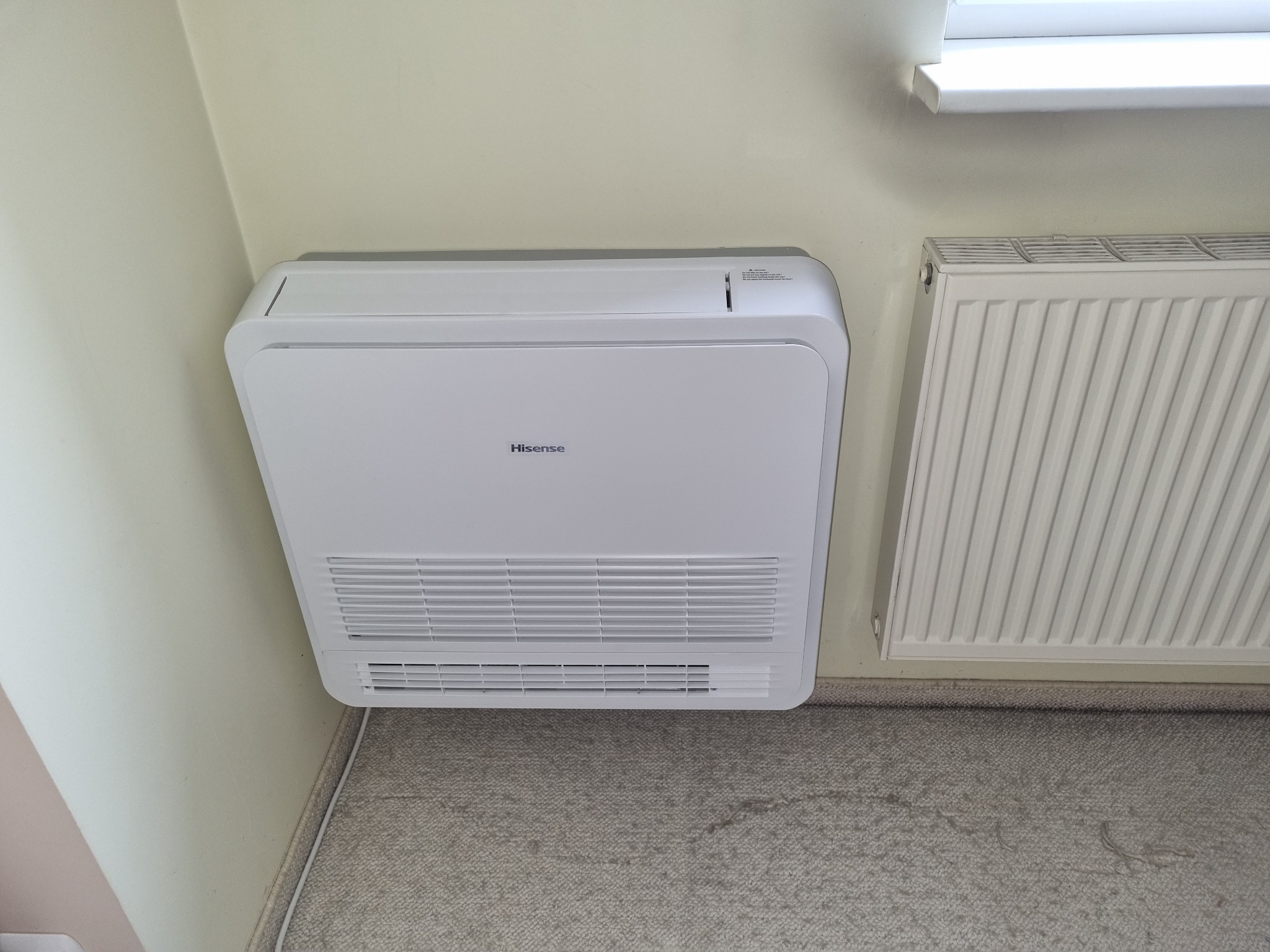 Nowoczesny, biały klimatyzator podłogowy marki Hisense zamontowany przy ścianie w jasnym pomieszczeniu, obok grzejnika.