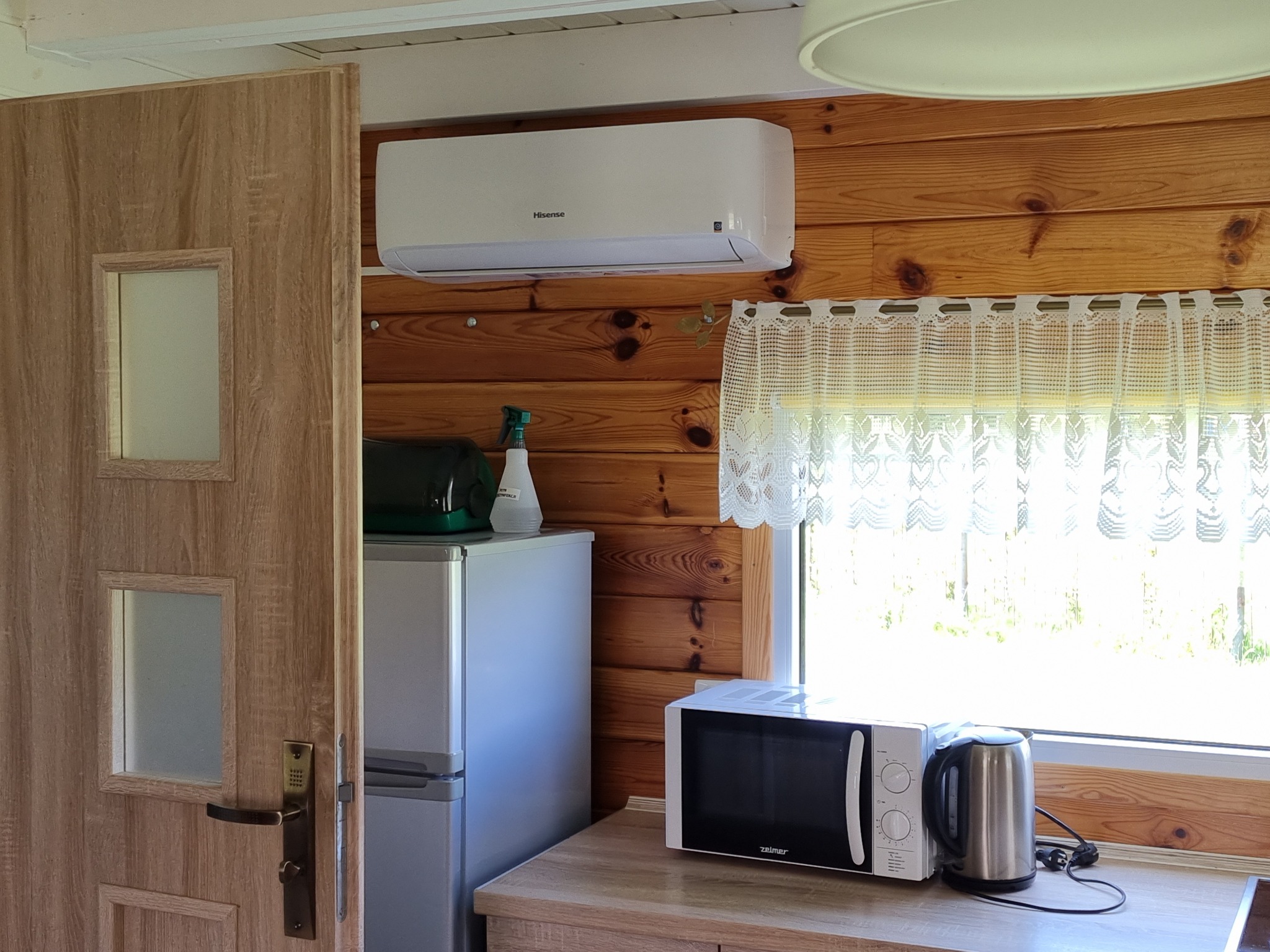 Biała klimatyzacja Hisense zamontowana na drewnianej ścianie w domku letniskowym, widoczny fragment okna z koronkową firanką, kuchenka mikrofalowa Zelmer i czajnik elektryczny na blacie