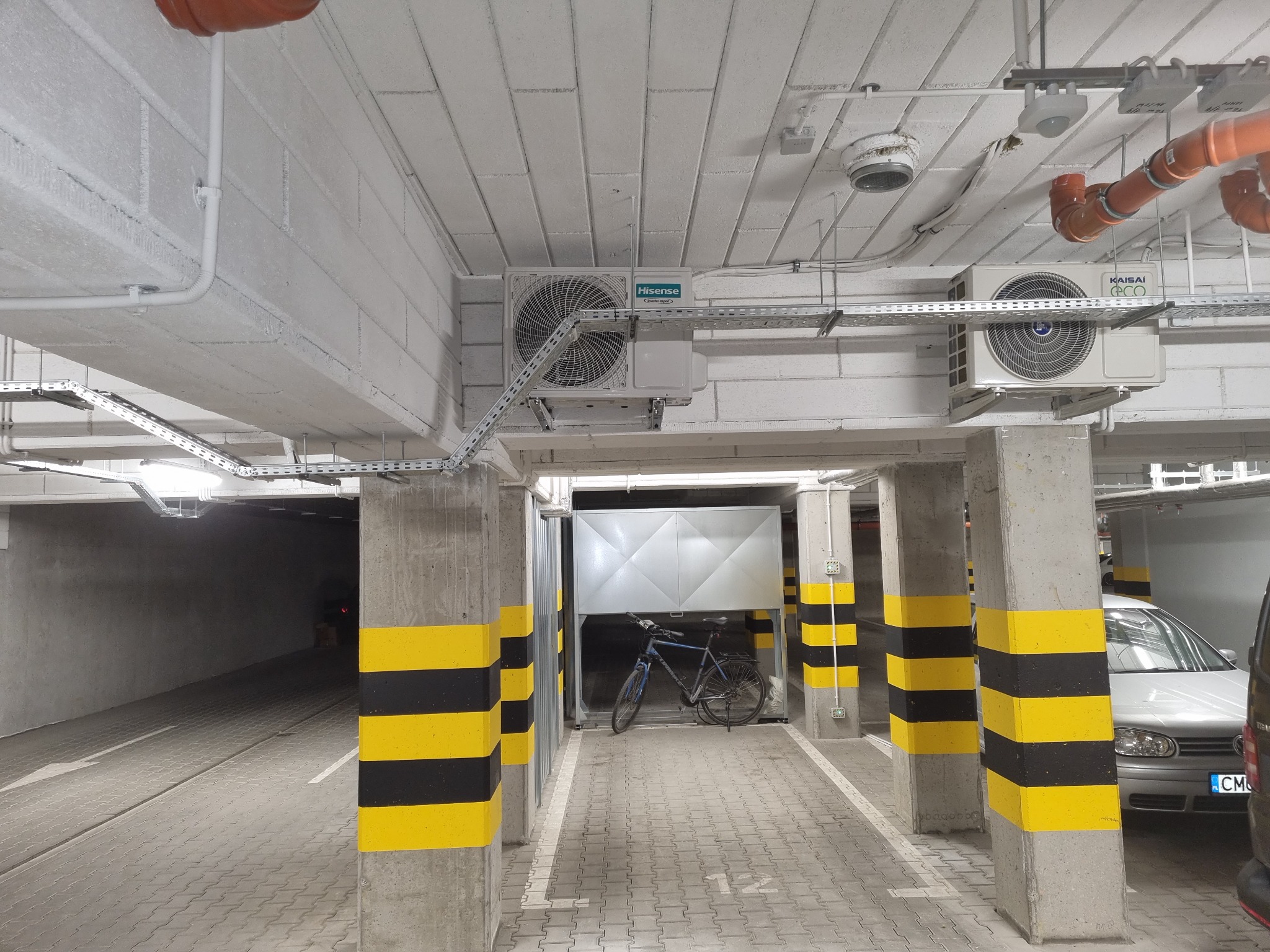 Dwa klimatyzatory zamontowane pod sufitem w garażu podziemnym, widoczne rury i kable instalacyjne.