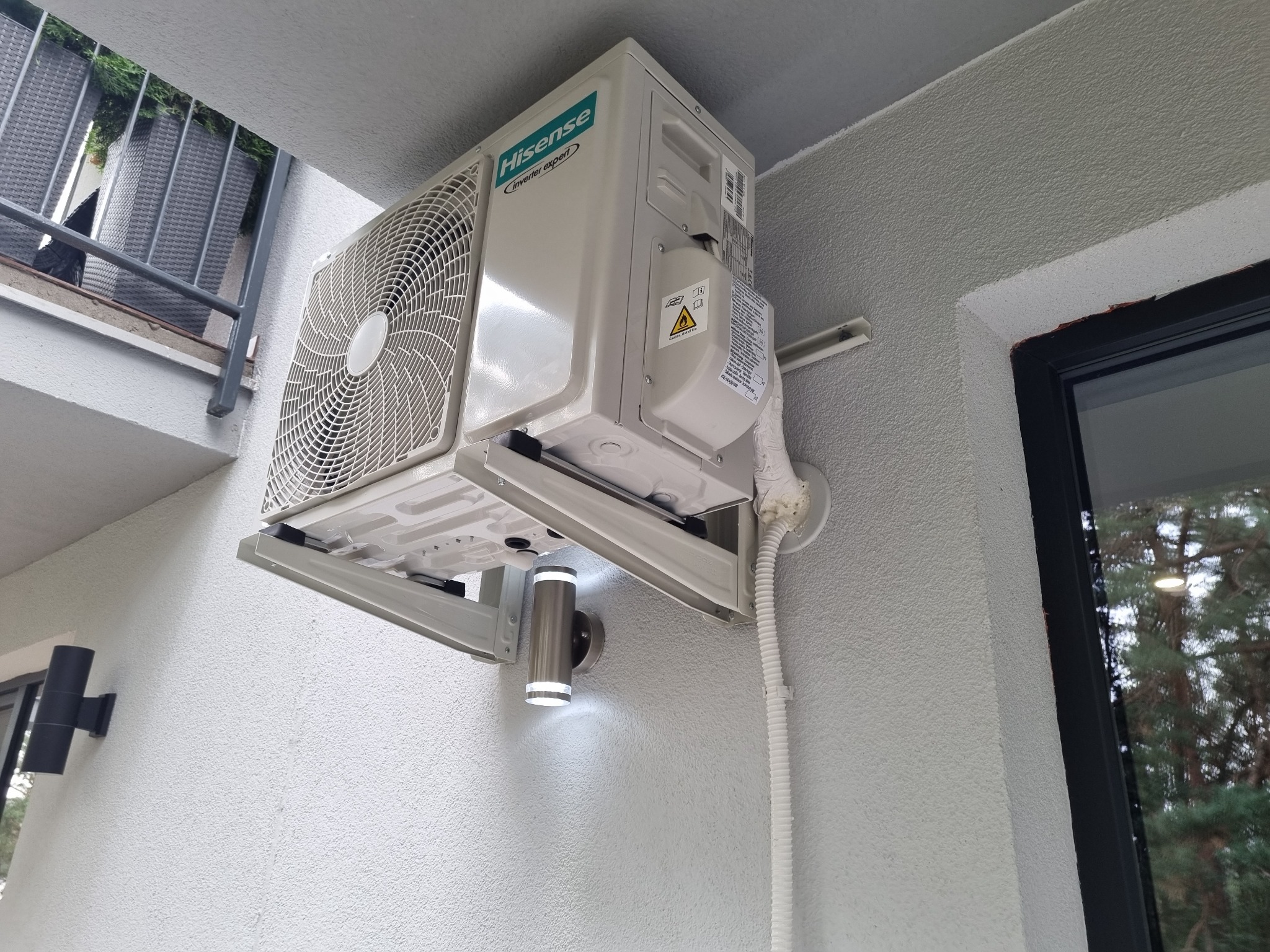 Biała jednostka zewnętrzna klimatyzatora Hisense Inverter Expert zamontowana na ścianie budynku z oknem i balkonem w tle.