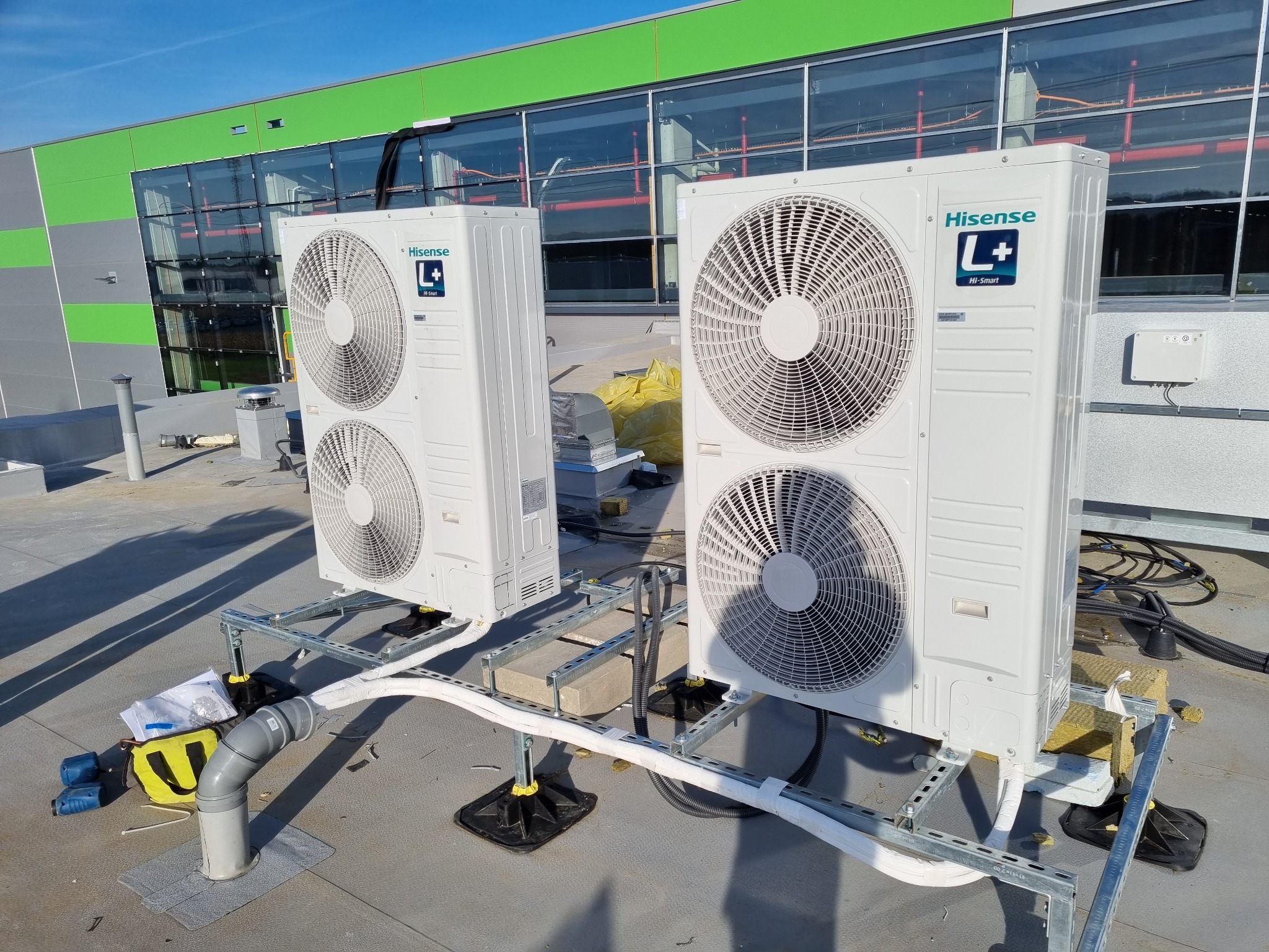 Dwie jednostki zewnętrzne klimatyzacji Hisense L+ Hi-Smart zamontowane na metalowej konstrukcji na dachu budynku z zielono-szarą elewacją, widoczna instalacja odprowadzająca skropliny i narzędzia...