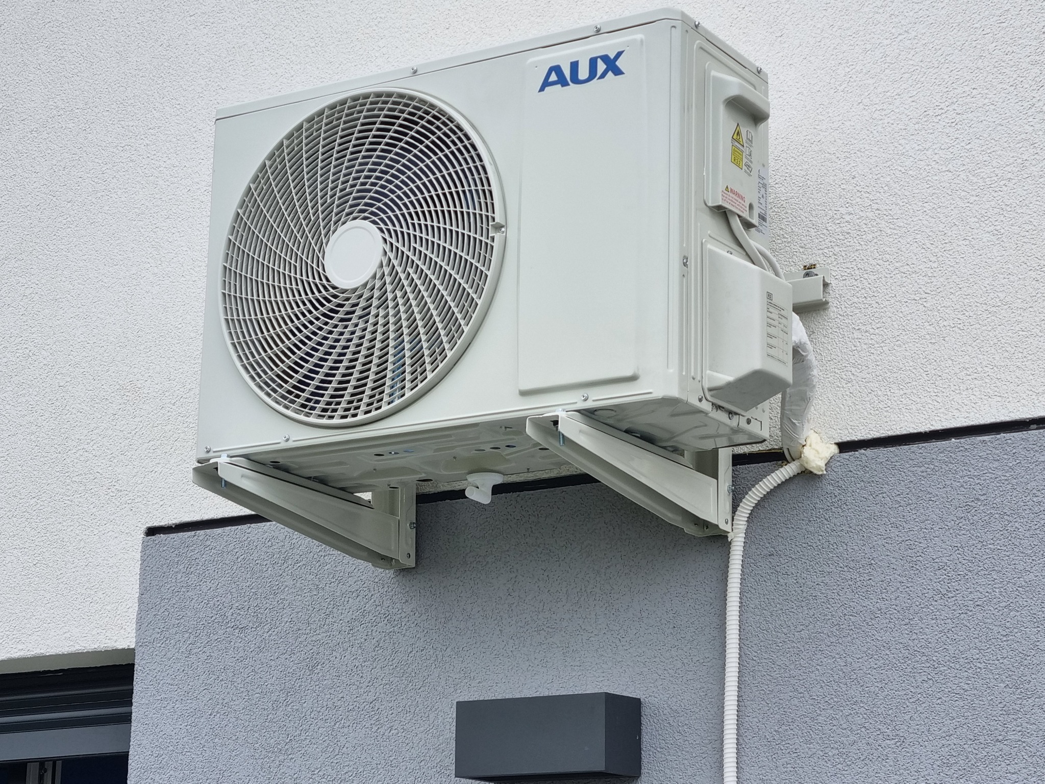 Zewnętrzna jednostka klimatyzatora marki AUX zamontowana na wspornikach na elewacji budynku z widocznymi elementami instalacji i uszczelnieniem pianką montażową.