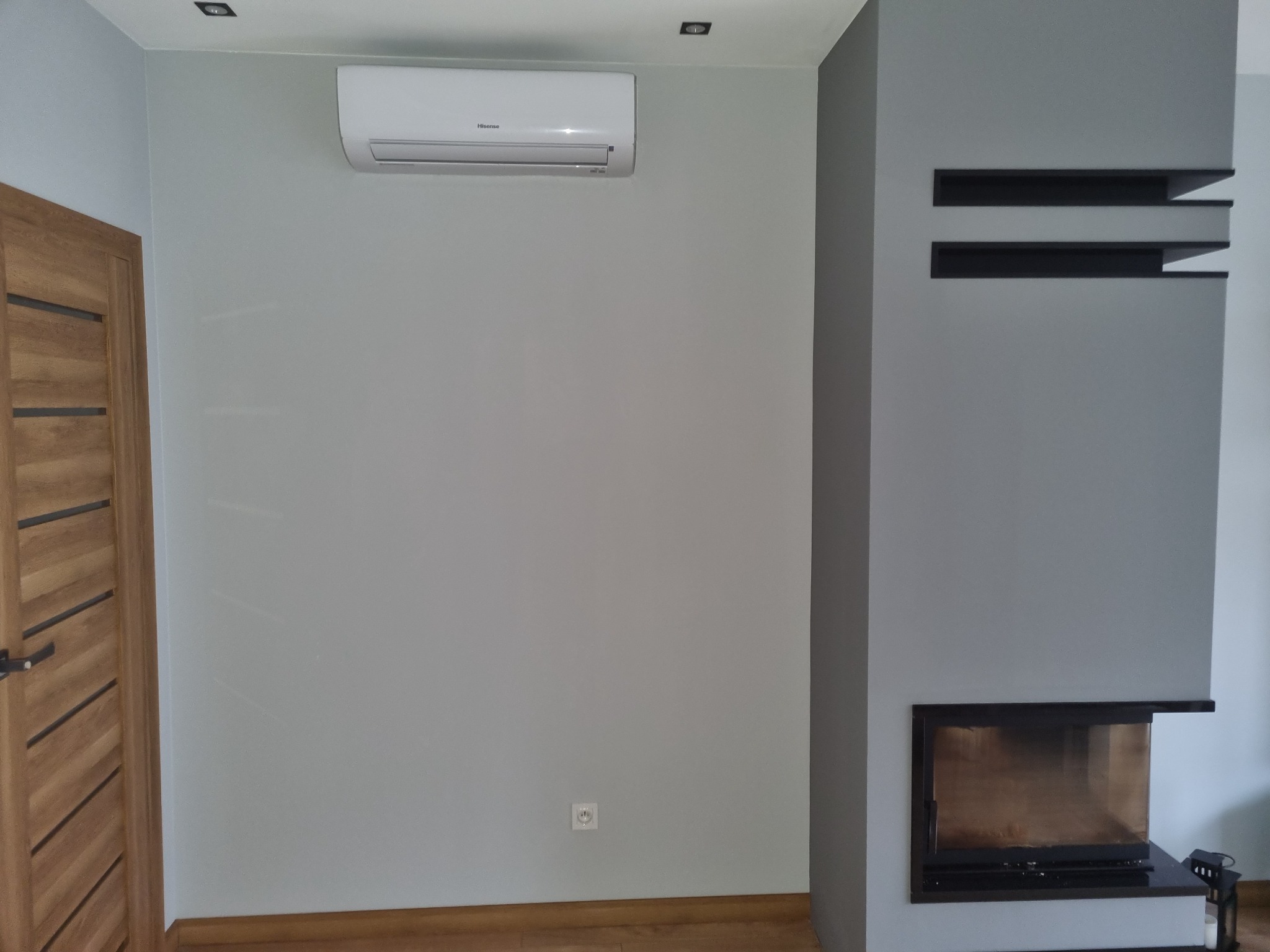Biała jednostka klimatyzacyjna Hisense zamontowana na szarej ścianie w jasnym pomieszczeniu, widoczny fragment drewnianych drzwi i kominek z półkami.