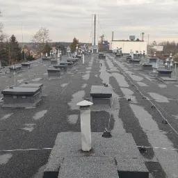 Płaski dach pokryty papą z licznymi kominami wentylacyjnymi i instalacją odgromową na tle zabudowań miejskich w pochmurny dzień.