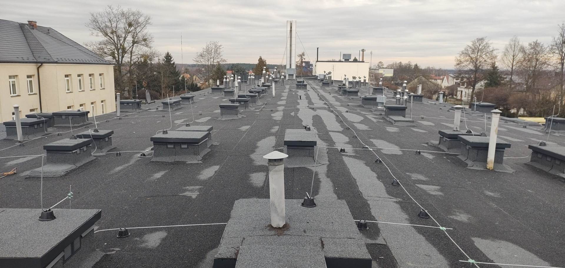 Płaski dach pokryty papą z licznymi kominami wentylacyjnymi i instalacją odgromową na tle zabudowań miejskich w pochmurny dzień.