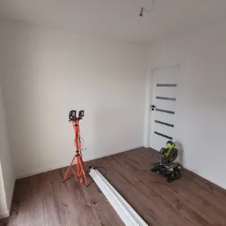 Pokój w trakcie wykańczania: lampa budowlana na trójnogu, listwy przypodłogowe, ukośnica Ryobi na podłodze z paneli, białe ściany i drzwi z poziomymi przeszkleniami.