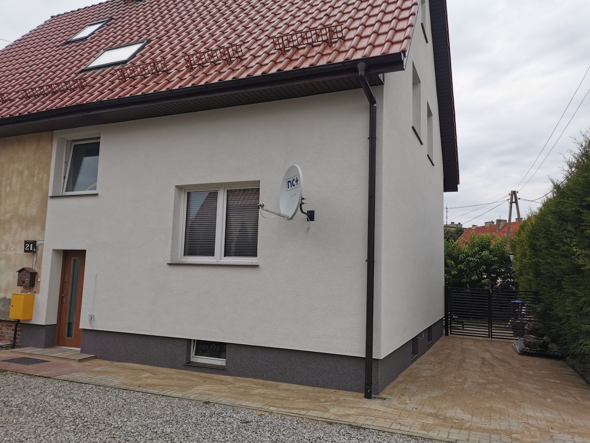 Odnowiona elewacja dwukondygnacyjnego domu z nową, czerwoną dachówką i brązowymi rynnami, satelitarna antena na bocznej ścianie, szara podmurówka, okno z żaluzjami.