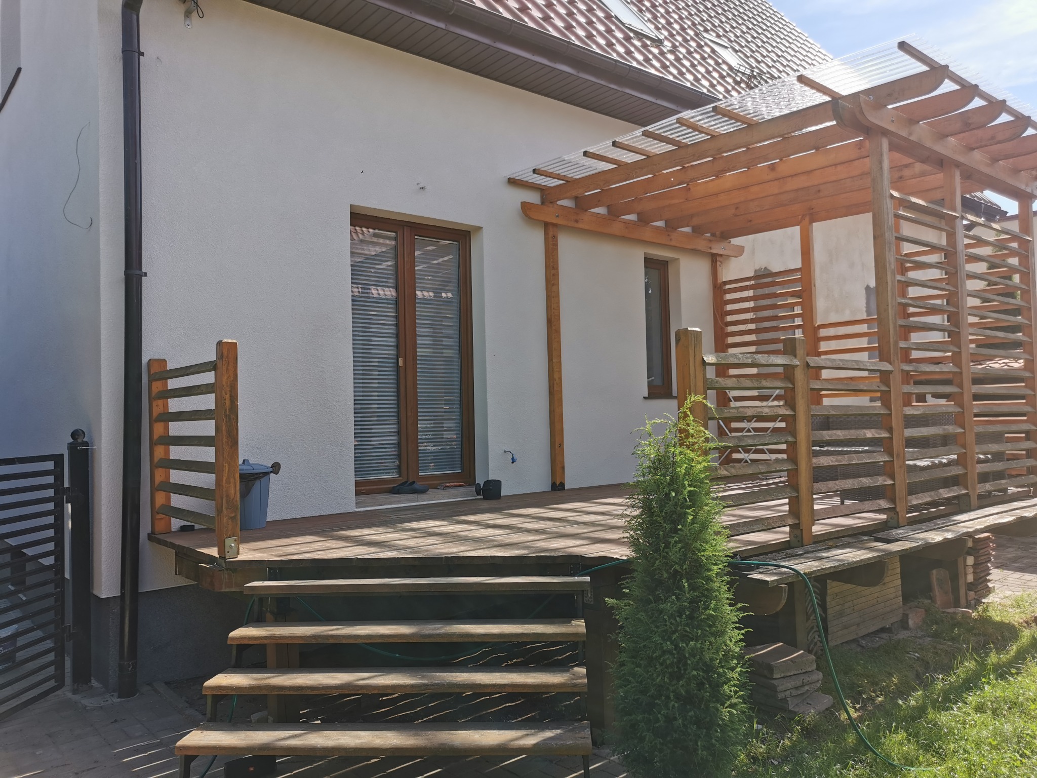 Drewniany taras z pergolą z przezroczystym zadaszeniem, schody z widocznym wężem ogrodowym, tuja obok schodów, biała ściana budynku z brązowymi oknami.