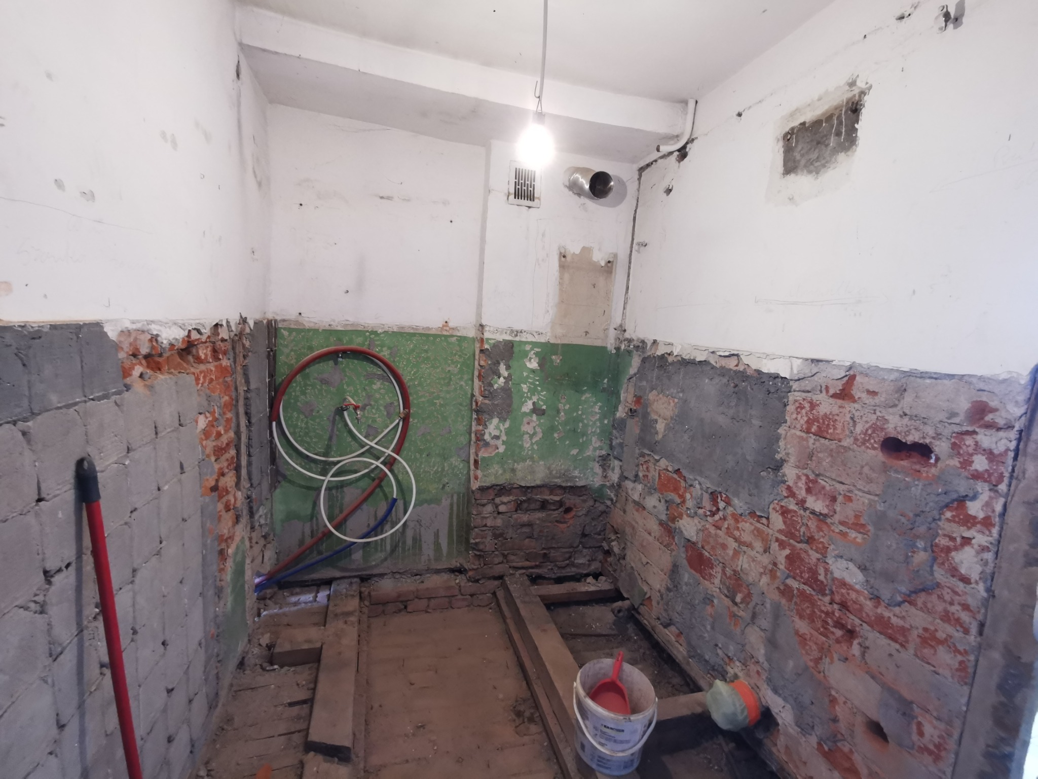 Wnętrze łazienki w trakcie demontażu starej instalacji: odsłonięte fragmenty ceglanej ściany, rury instalacji wodnej, resztki podłogi i widoczne ślady po starych płytkach na ścianach.